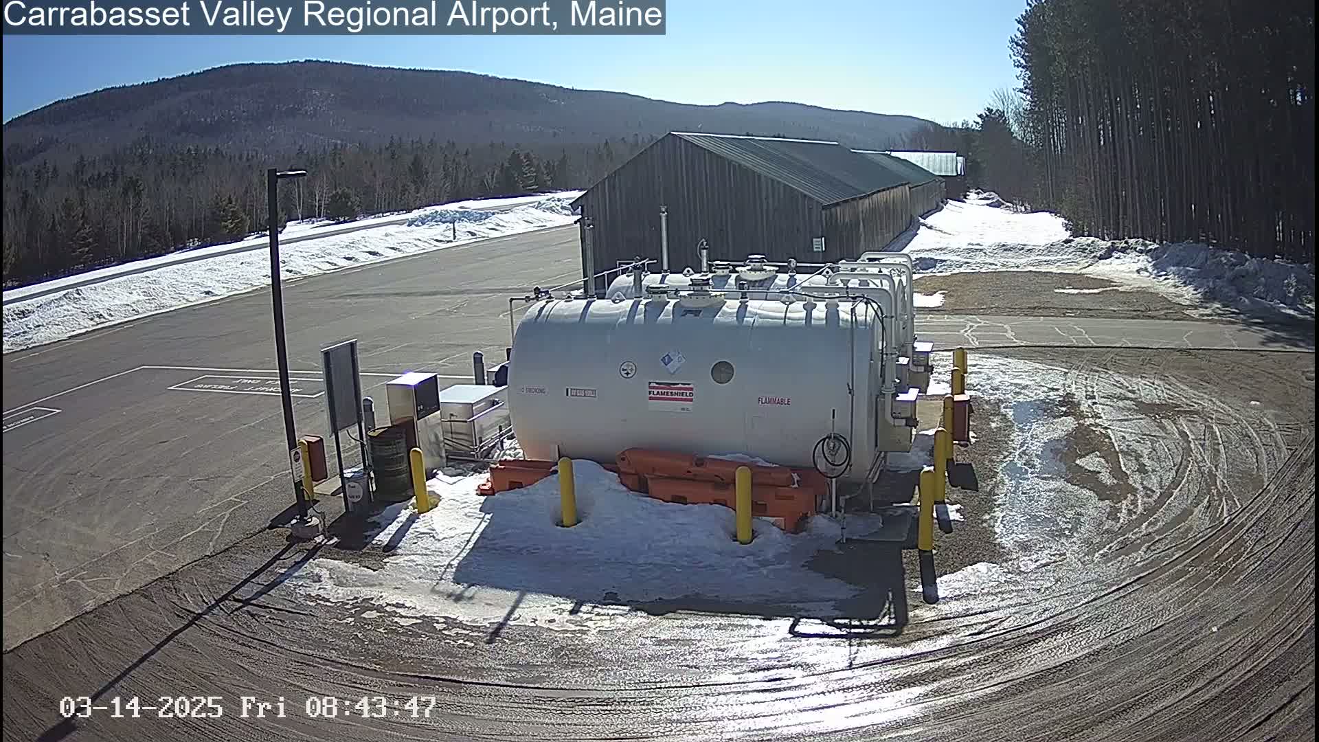 A sunny day reveals several large, white, cylindrical fuel tanks surrounded by patchy snow in a parking lot next to a long, dark-colored building and a snow-covered, wooded hillside.