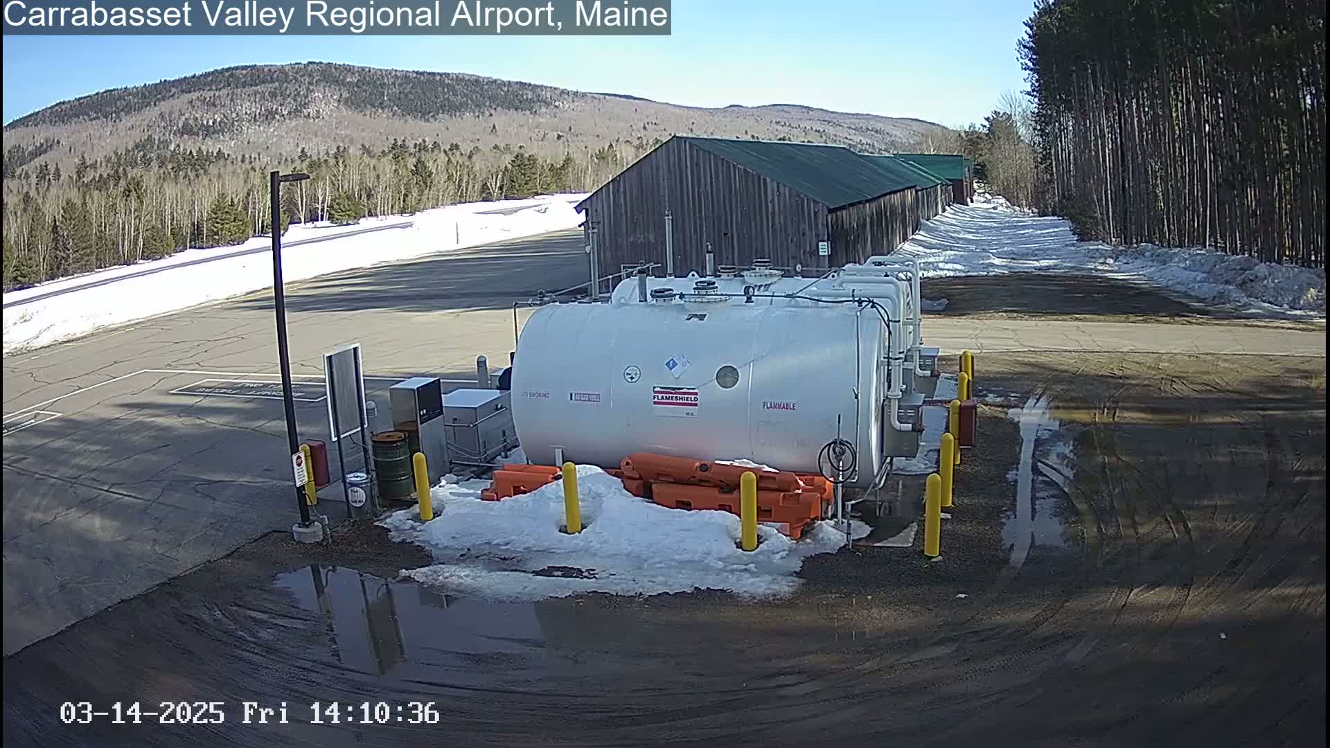 A large white fuel tank sits in a snow-covered parking lot next to a long, low building under a clear, sunny sky.