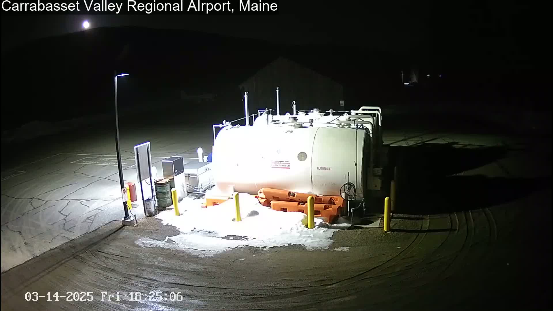 A large white fuel tank sits in a snow-dusted, nighttime airport parking lot, illuminated by artificial light.