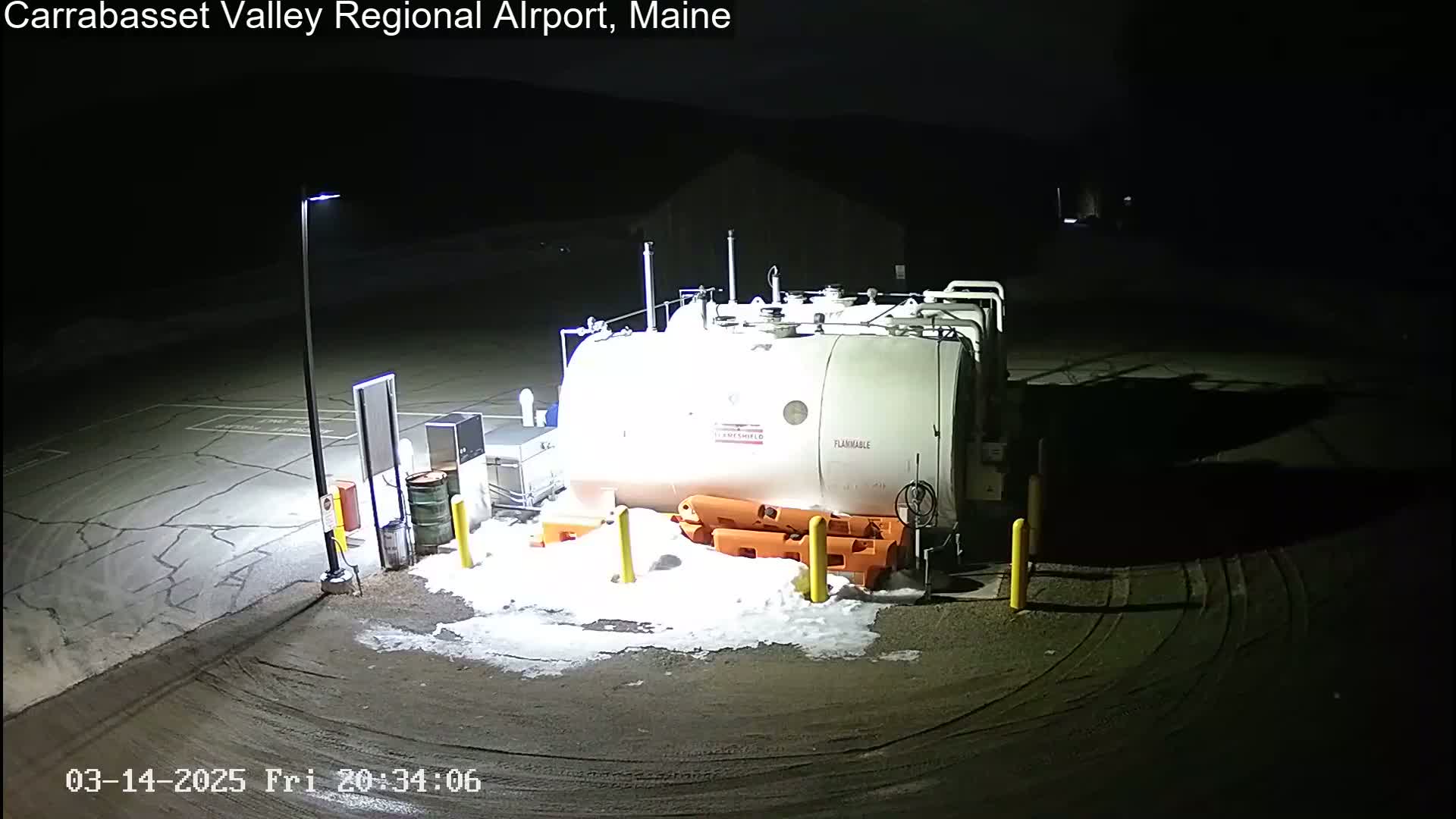 A large white fuel tank sits in a snow-dusted parking lot at night, illuminated by a single light pole.