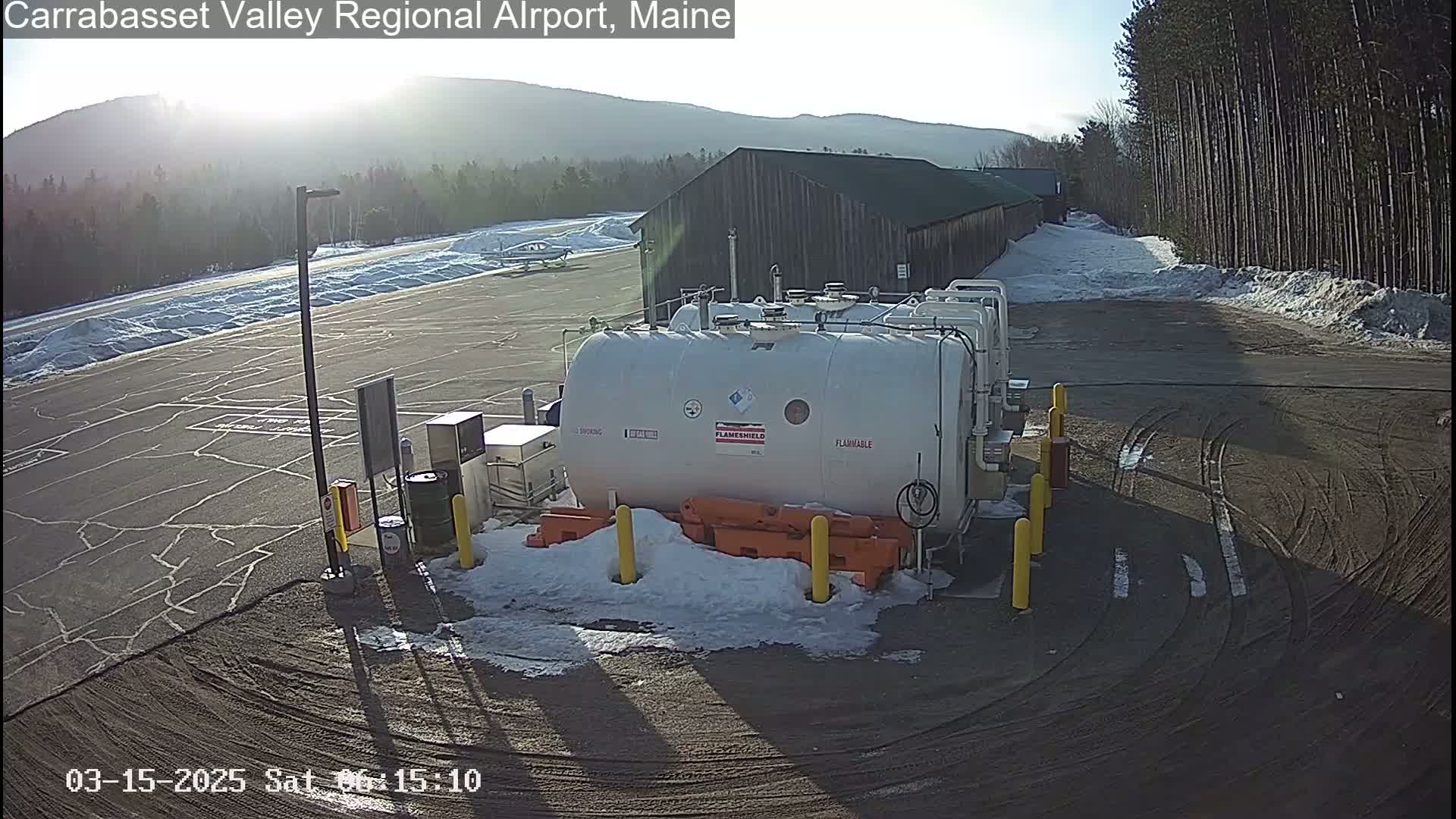 A large, white, flammable fuel tank sits in a snowy, partially paved lot near a hangar-like building under a bright, sunny sky.
