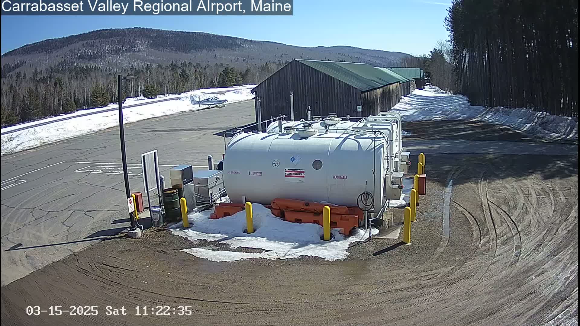 A large, white fuel tank sits in a snowy airport parking lot next to a small airplane and a wooden building, under a sunny sky.