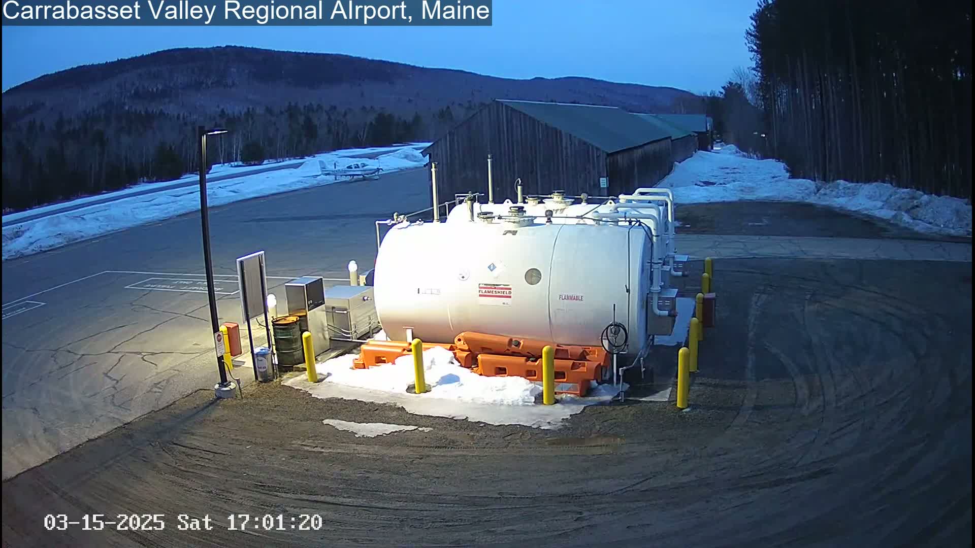 A large white fuel tank sits in a snowy airport parking lot at dusk, with a small airplane visible in the background.