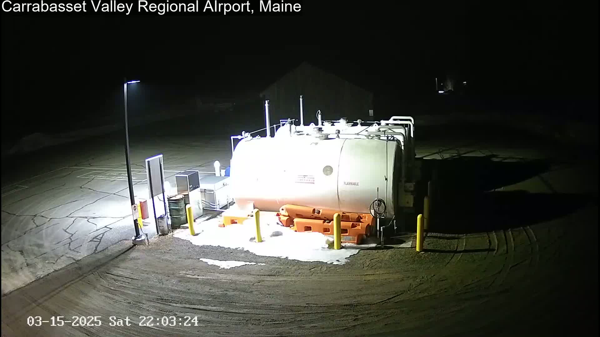 A large, light-colored fuel tank sits in a dimly lit, snow-dusted airport parking lot at night.