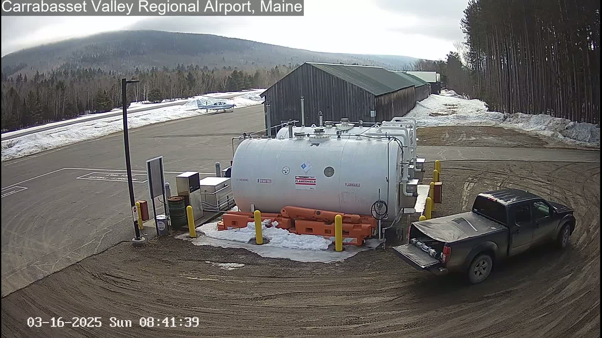 A dark-colored pickup truck is parked near several large white fuel tanks at an airport on a partly cloudy day with snow on the ground.