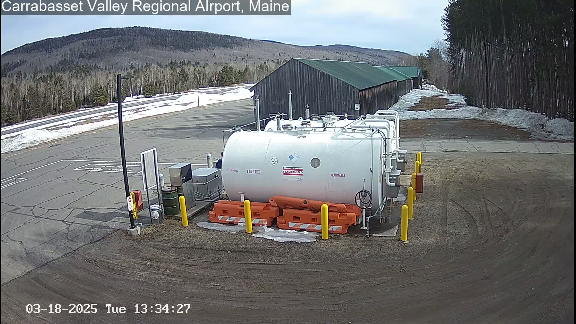 A large white fuel tank sits in a mostly paved area next to a long, dark-colored building, with snow on the ground and trees in the background under a mostly clear sky.