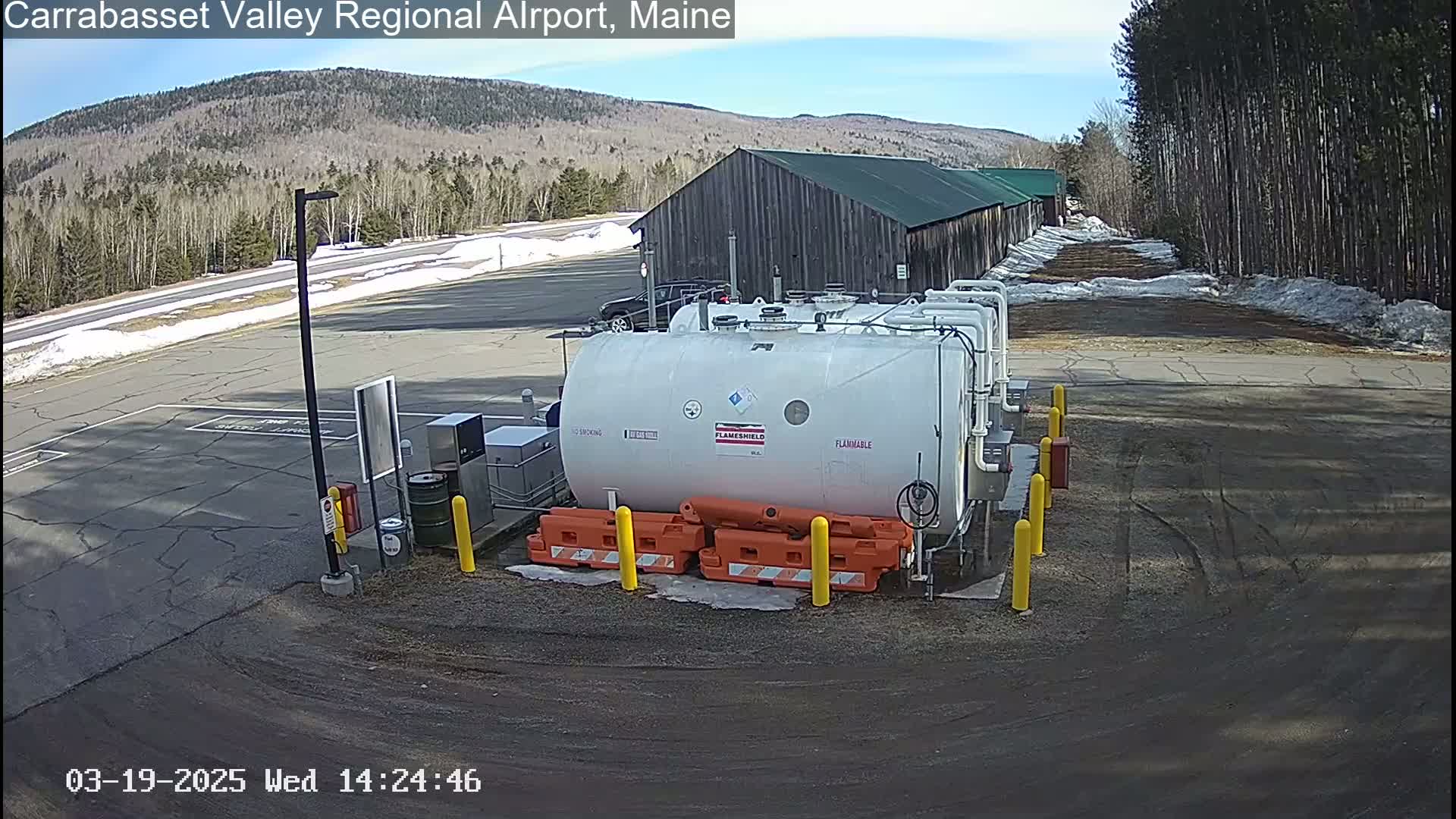 A large white fuel tank sits in a paved area next to a long, low wooden building, with a mostly clear sky and patches of snow on the ground.