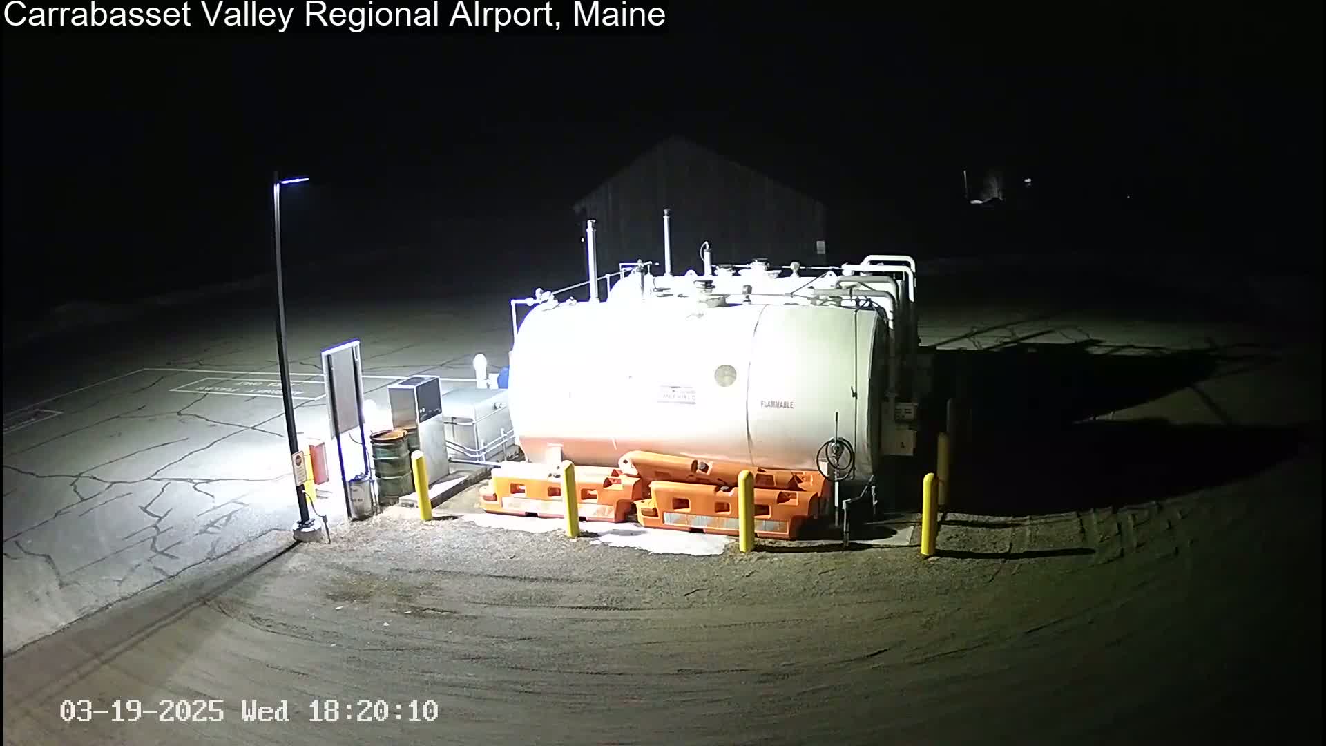 A large, white fuel tank sits in a dark, empty, asphalt lot at night, illuminated by a single lamppost.