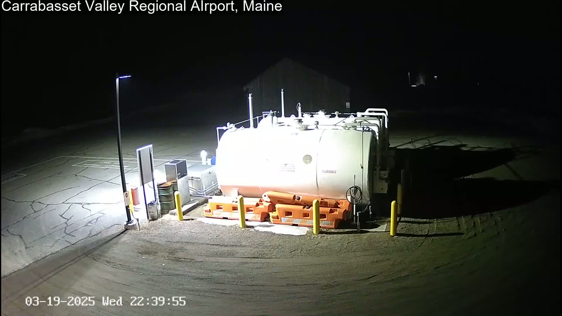 A large, cylindrical fuel tank sits in a dark, nighttime airport lot, illuminated by a single lamppost.