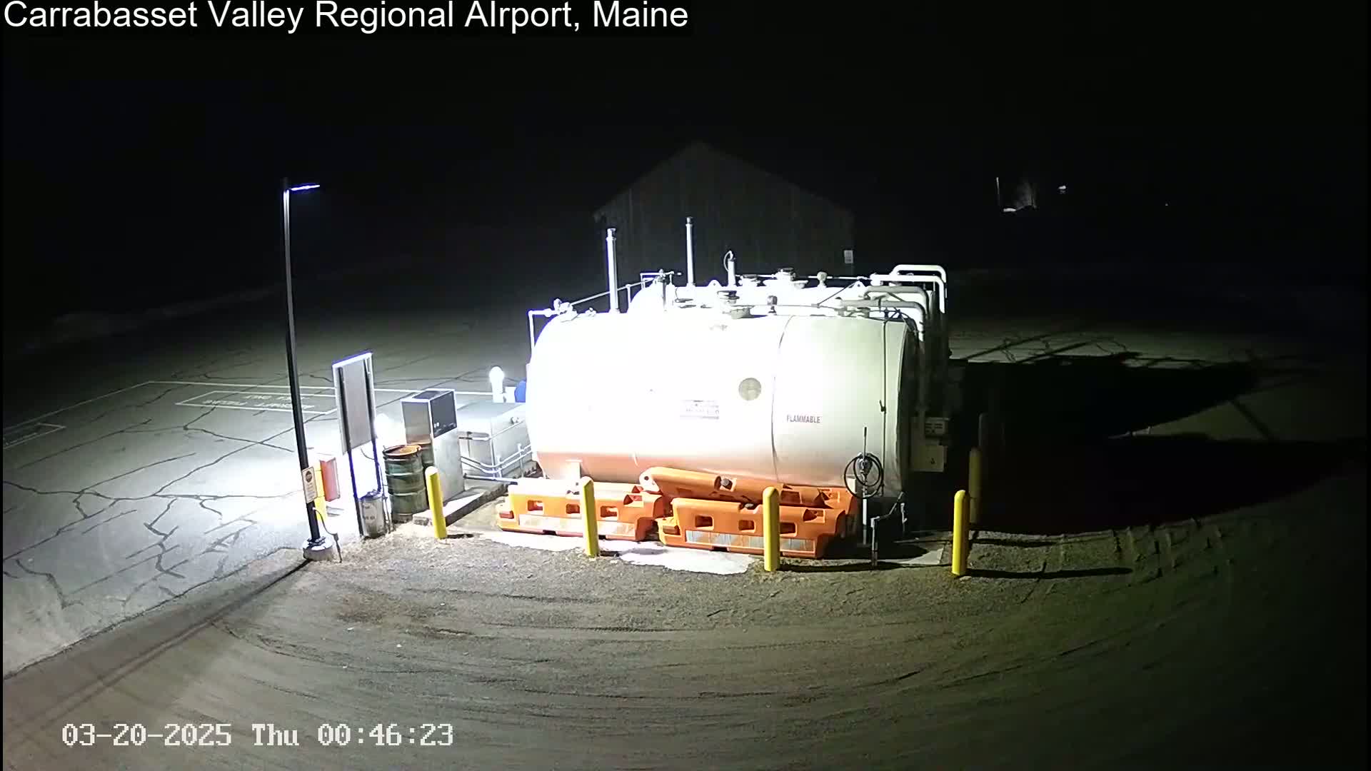 A large, white fuel tank sits in a dark, empty airport lot at night, illuminated by a single lamppost.
