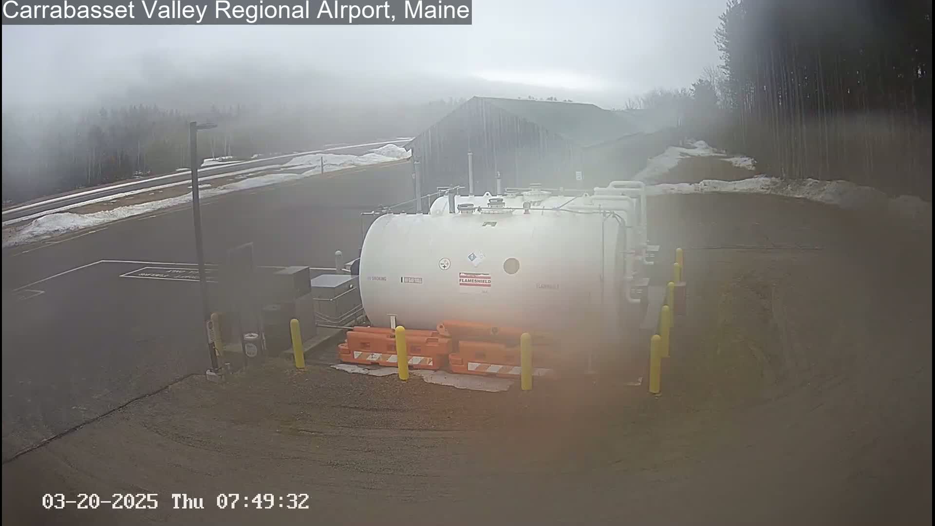 A large white fuel tank sits in a foggy parking lot near a building, with patches of snow visible on the ground.