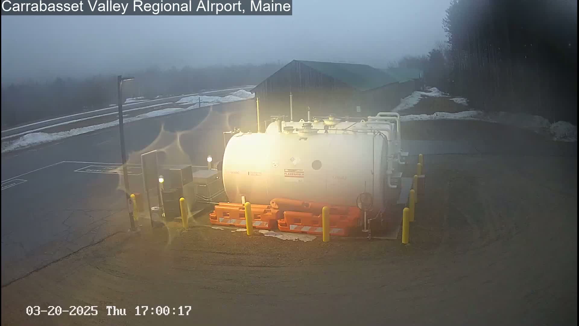 A large white tank surrounded by orange barriers sits in a foggy, dimly lit outdoor parking lot.