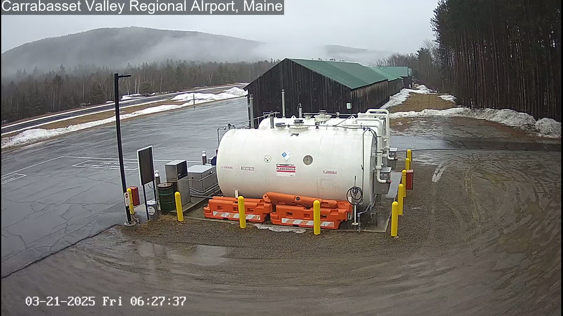 A large white flammable liquid storage tank sits in a wet, mostly empty parking lot next to a dark-colored building under an overcast sky.