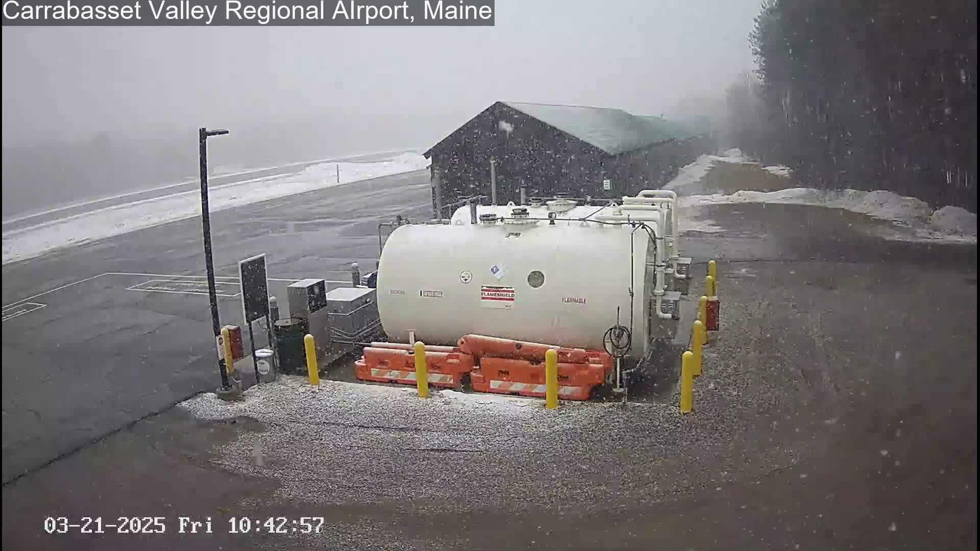 A large white tank sits in a snow-covered airport parking lot during a light snowfall.