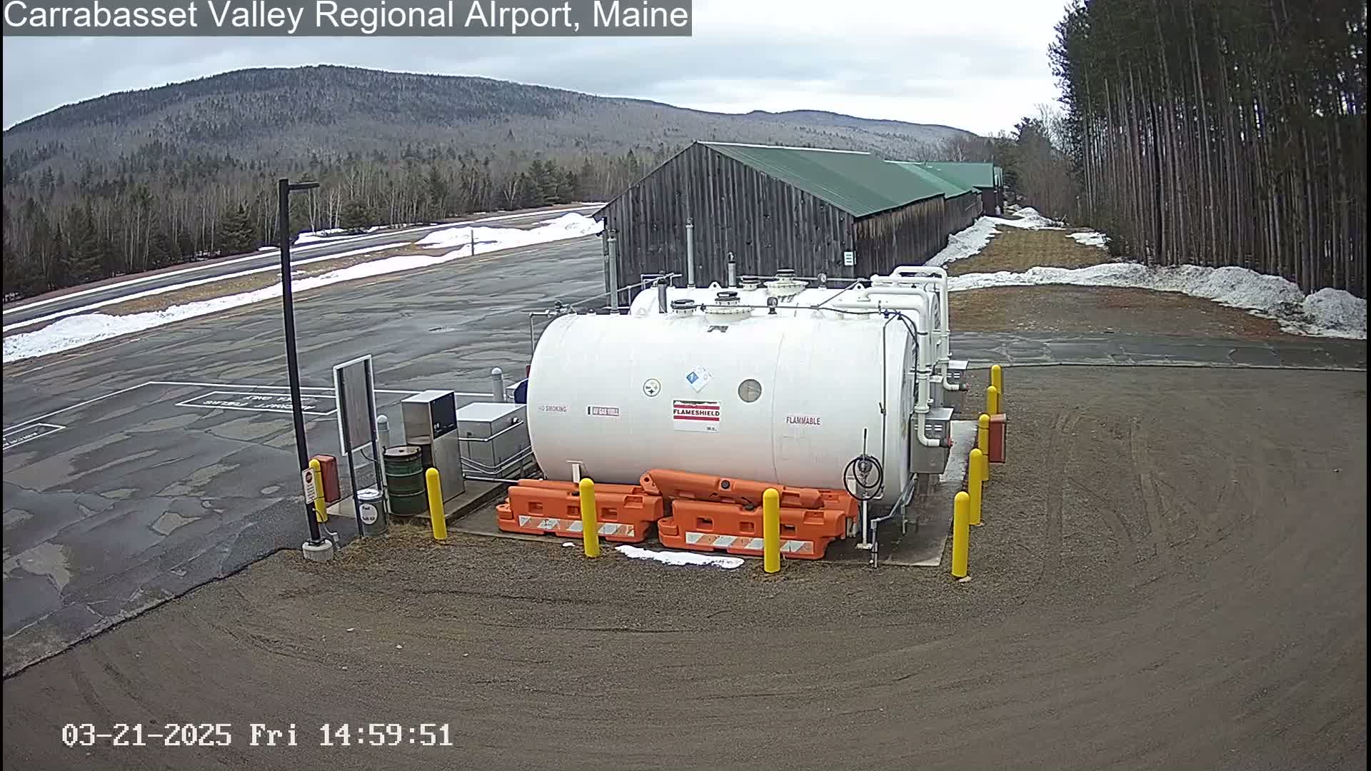 A large white fuel tank sits in a mostly empty parking lot, with patches of snow on the ground, in front of a long, low building and a treeline under a cloudy sky.
