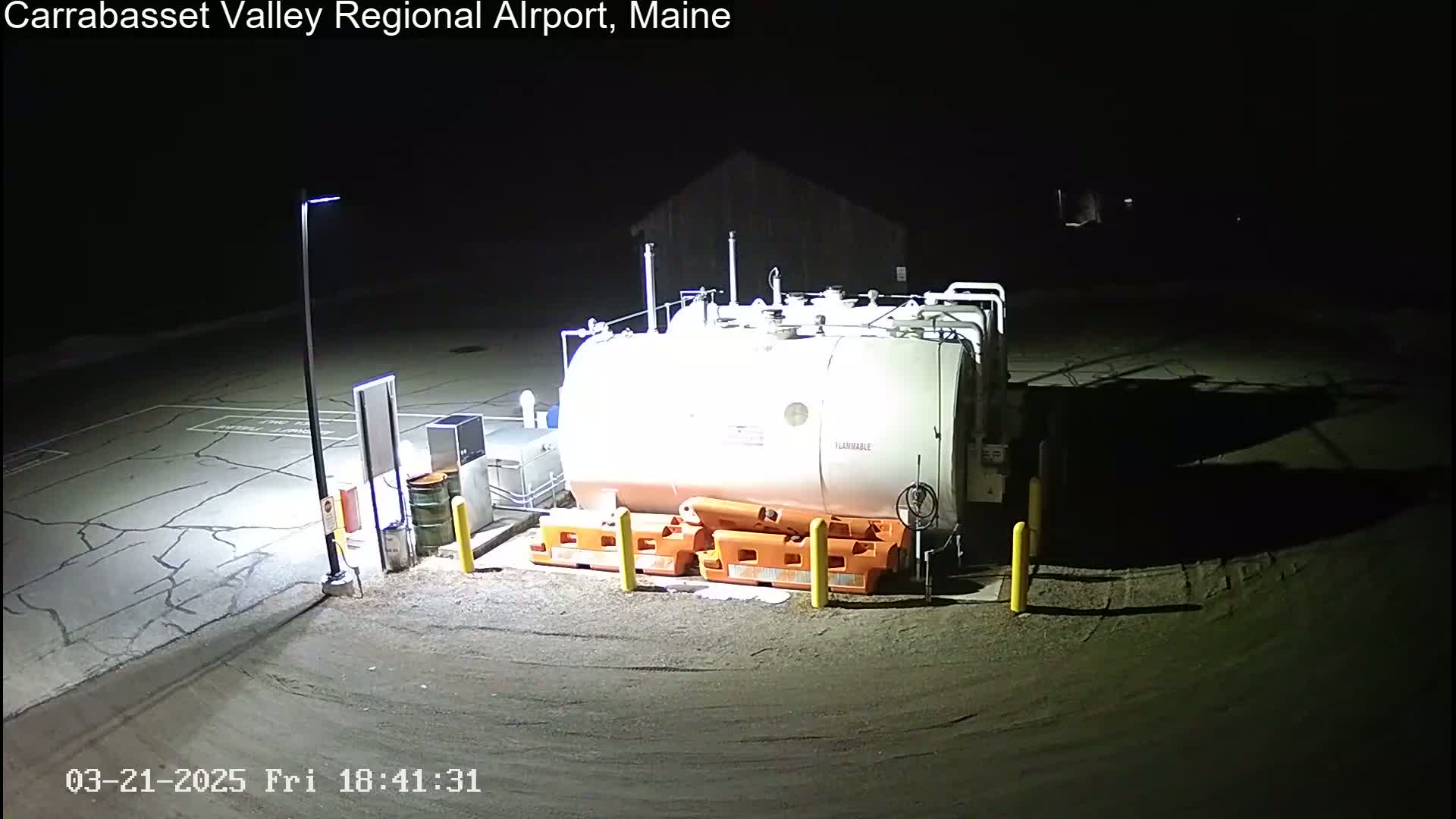 At night, a large white fuel tank sits in a paved area, surrounded by orange safety barriers and illuminated by artificial light.
