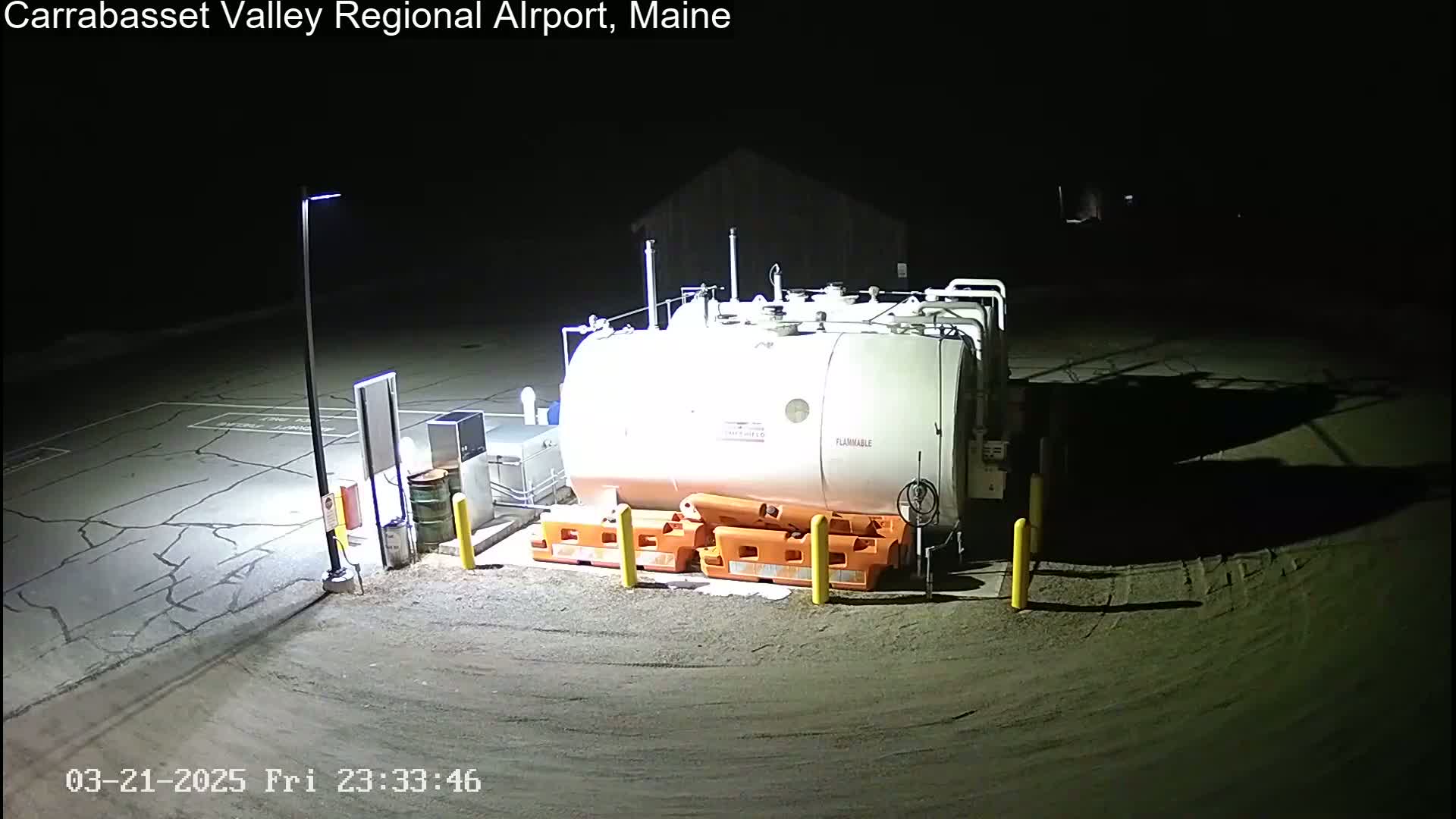 A large white fuel tank sits in a dimly lit, cracked asphalt area at night, surrounded by orange safety barriers and yellow posts.