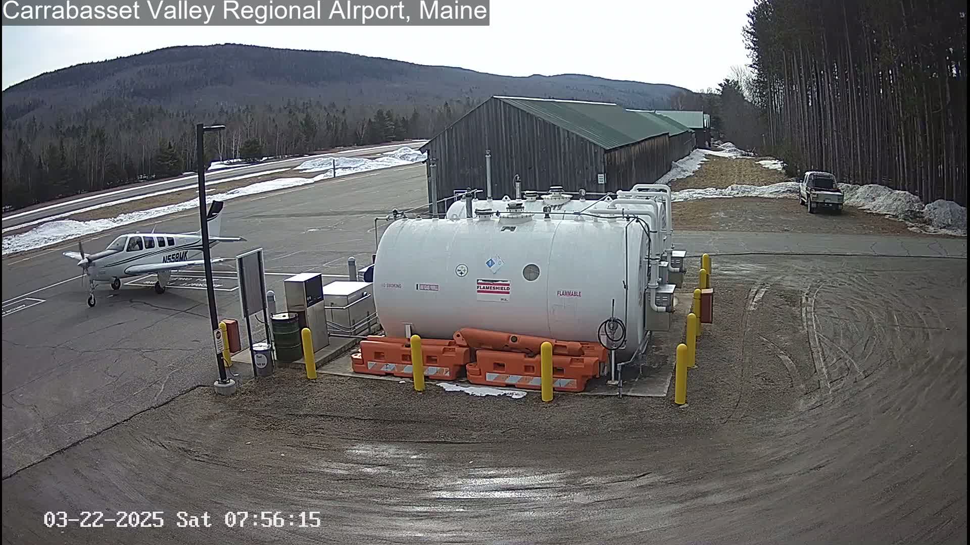 A small airplane is parked near several large fuel tanks at an airport on a partly cloudy day with some snow on the ground.