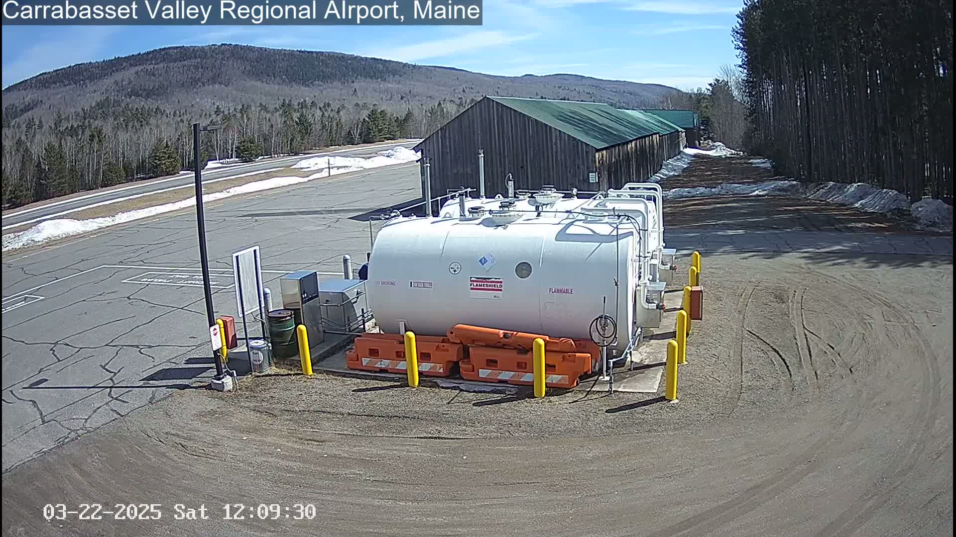 A large white fuel tank sits in a paved area of an airport, surrounded by snow-dusted ground and trees under a sunny sky.