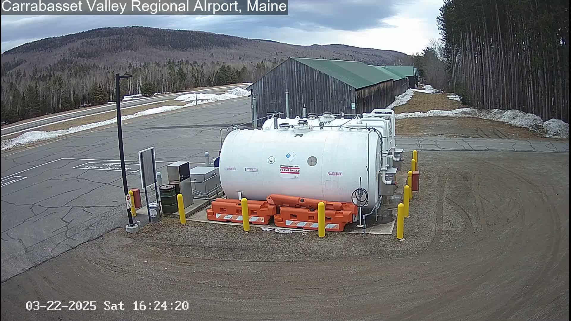 A large white fuel tank sits in a gravel parking lot near a wooded area under a partly cloudy sky.