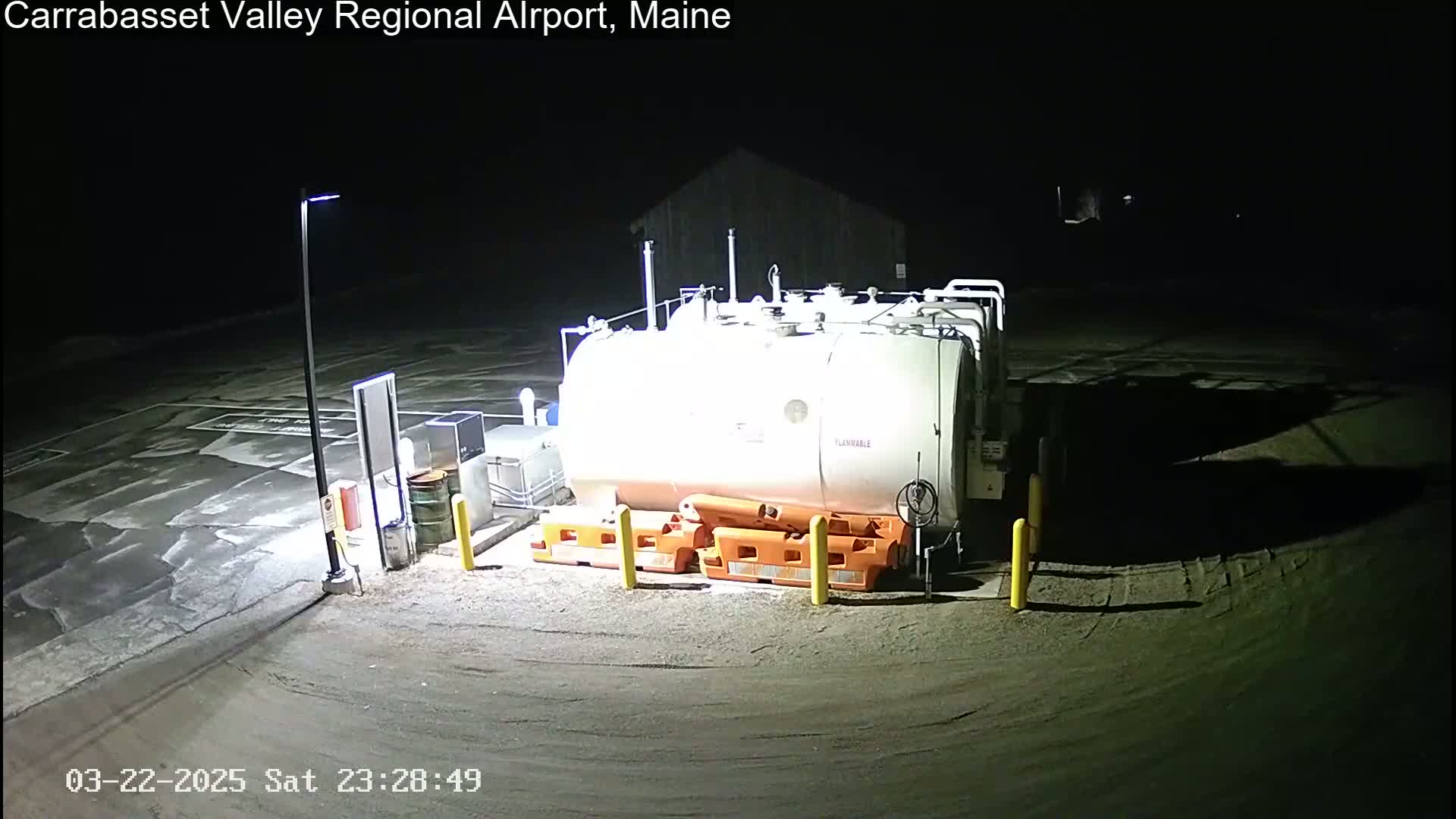 A large white fuel tank sits in a dark, nighttime airport lot, illuminated by a single light pole.
