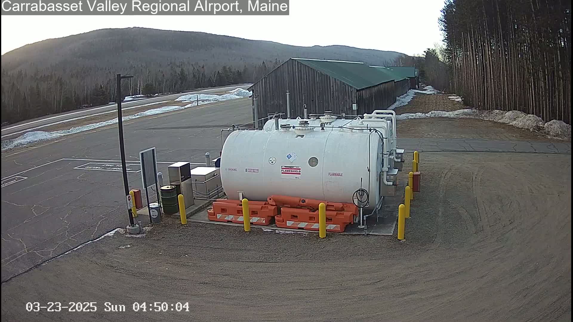 A large white fuel tank sits in a mostly empty asphalt lot next to a long, dark brown building, under a partly cloudy sky.