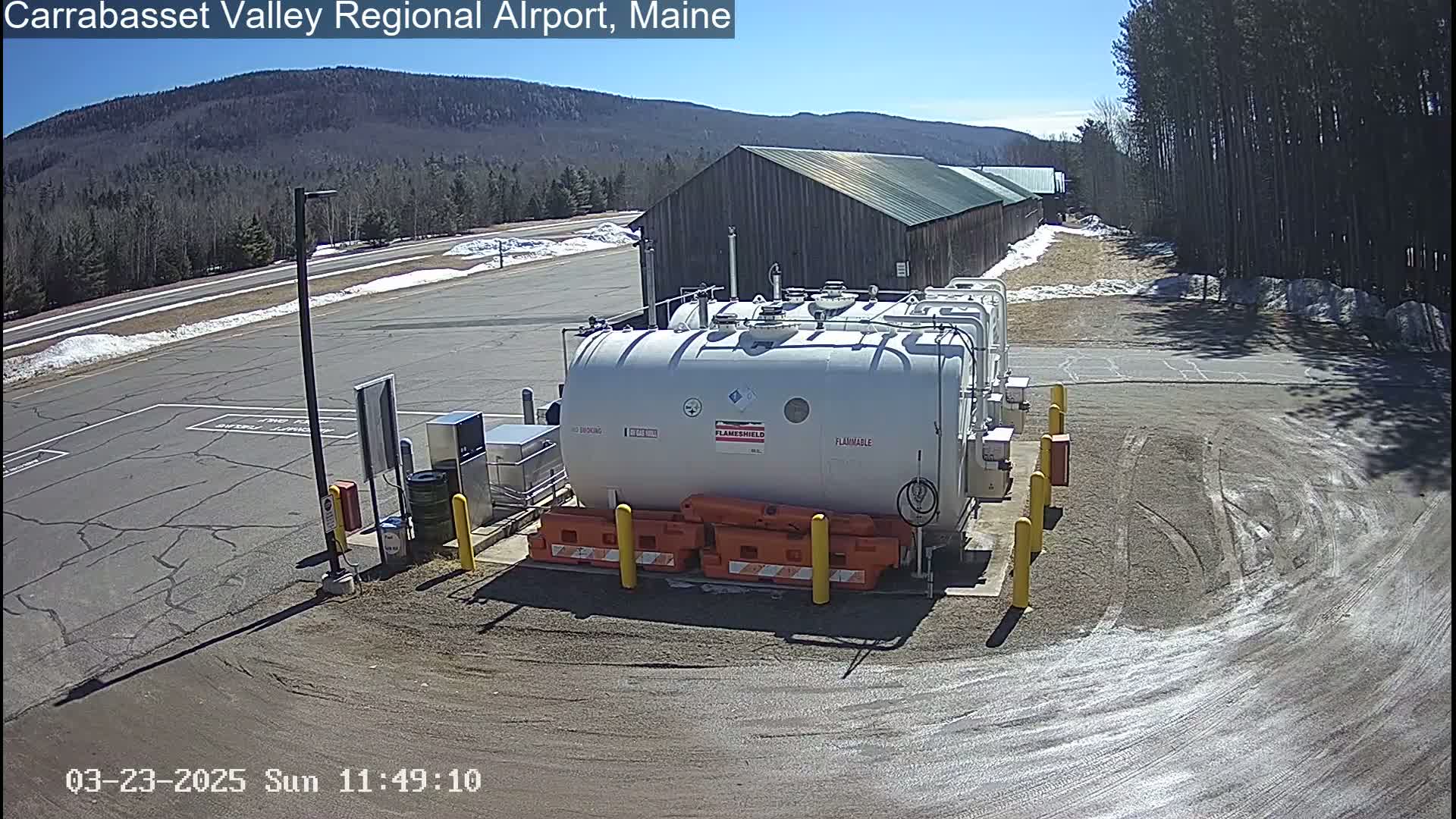 A large white fuel tank sits in a gravel area of an airport on a sunny day, with a long brown building and distant mountains visible in the background.