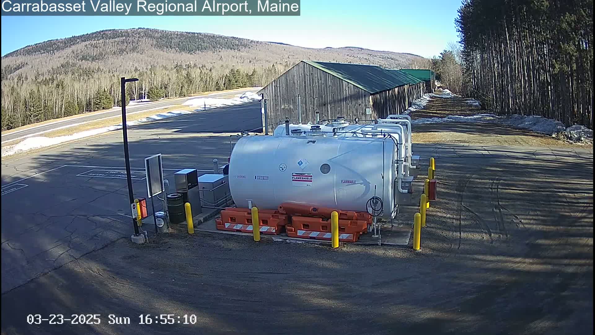 A large, light-grey fuel tank sits in a paved area of an airport on a sunny day, with a nearby building and forested hills in the background.