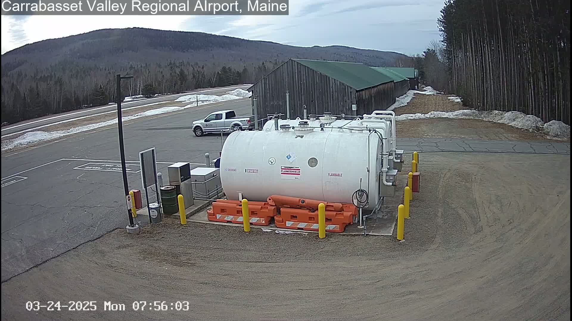 A large white fuel tank sits in a gravel lot near a pickup truck and a long wooden building, with a partly snowy landscape and distant mountains visible under a mostly cloudy sky.
