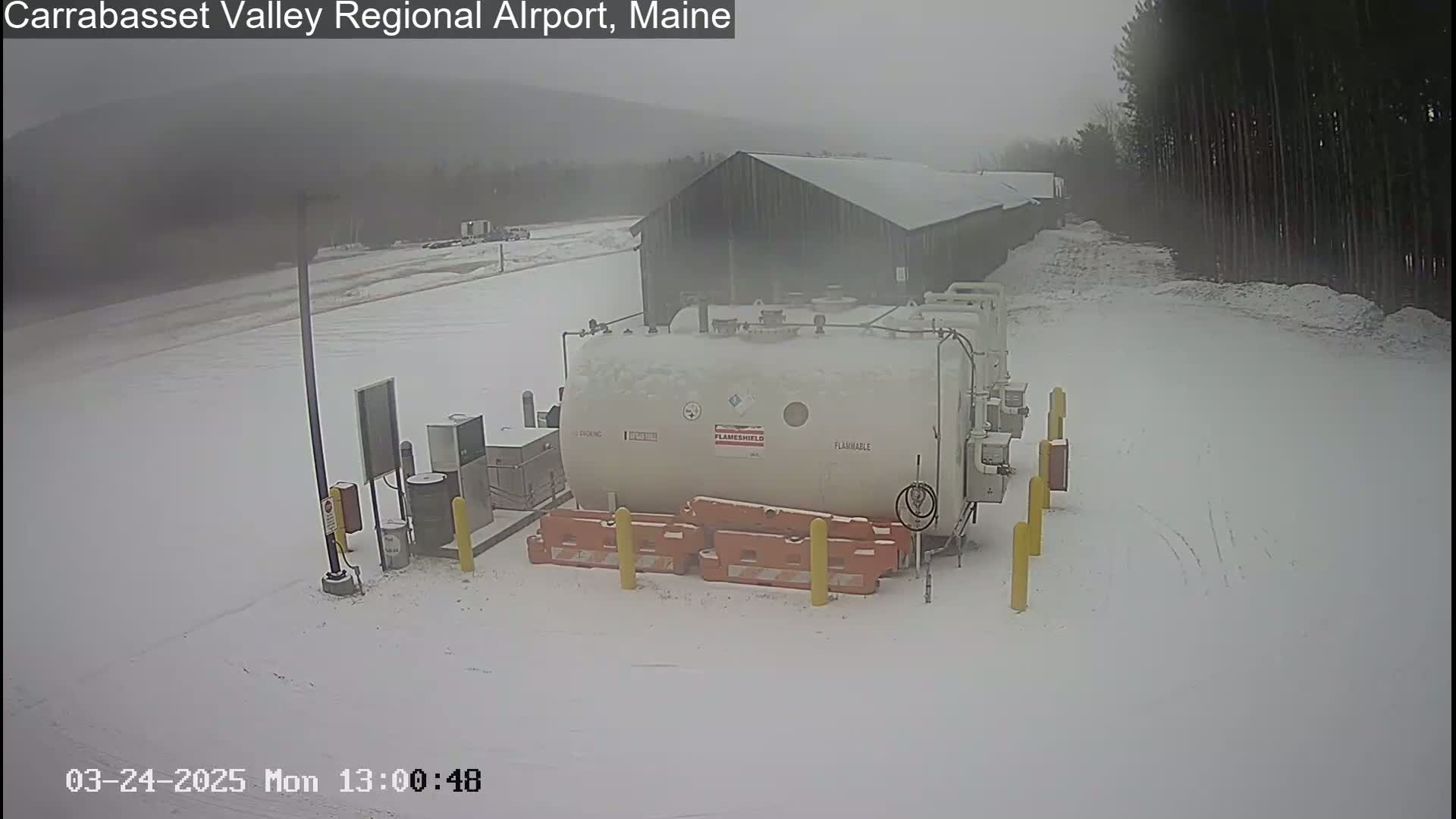 A large, white fuel tank sits in a snow-covered area next to a dark green building, under an overcast sky.
