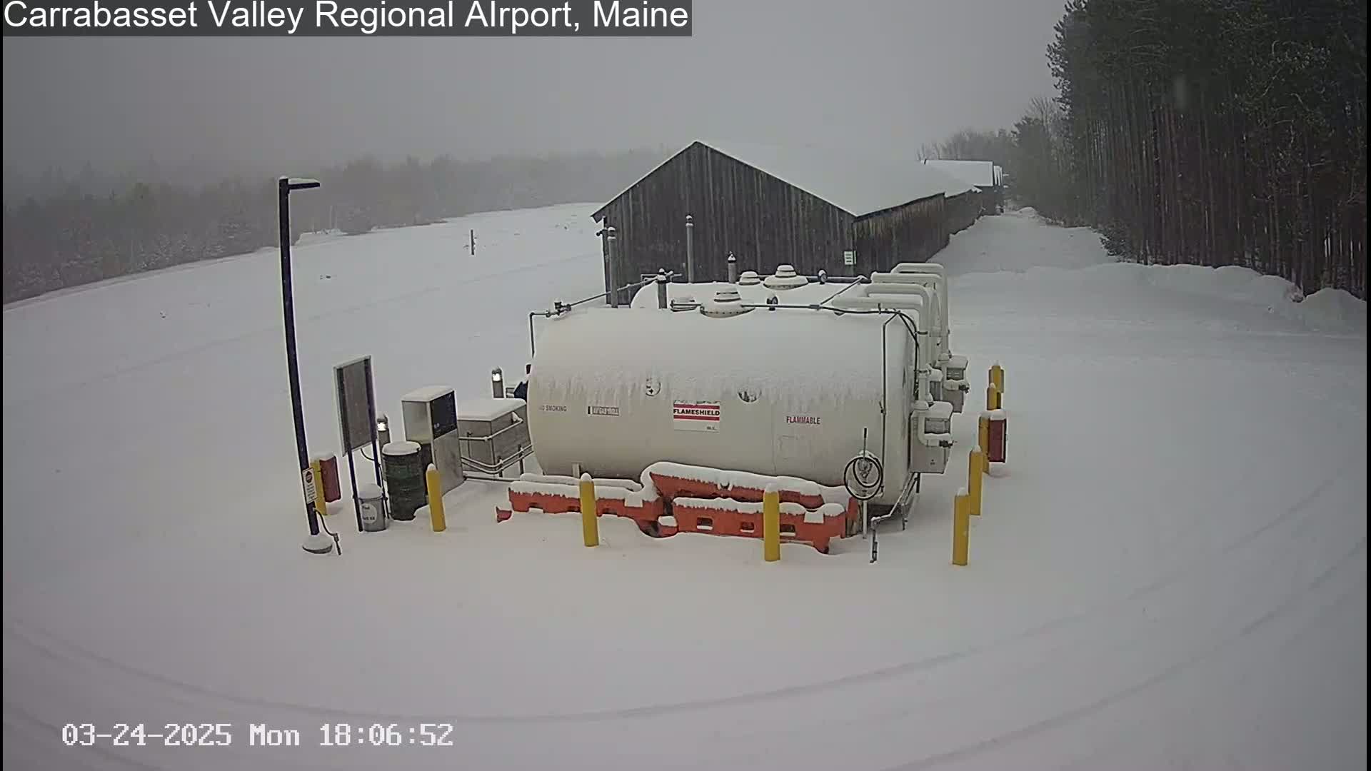 A snow-covered fuel tank sits in a snowy airfield near a dark-colored building, under a gray, overcast sky.