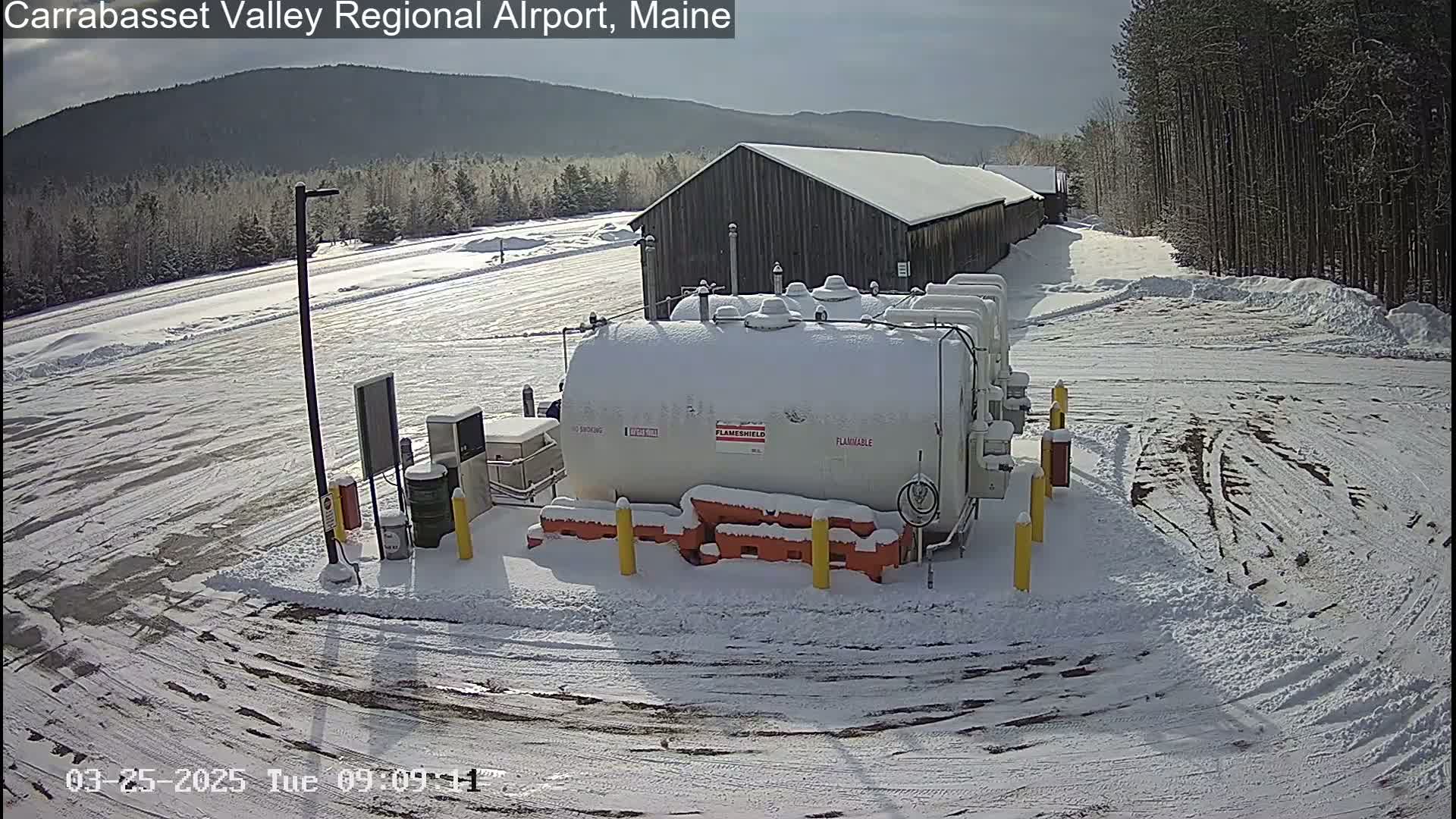 A snowy airfield scene shows several large white fuel tanks near a long, dark building under a bright, sunny sky.