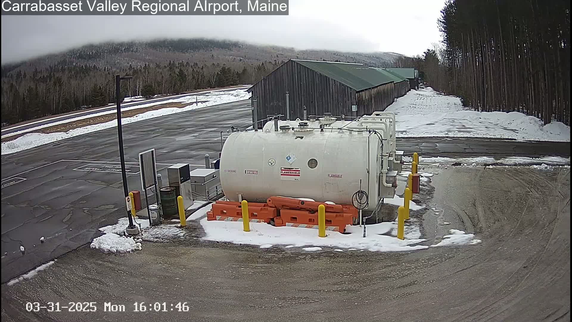 A large white flammable liquid storage tank sits in a snowy parking lot near a long wooden building under an overcast sky.