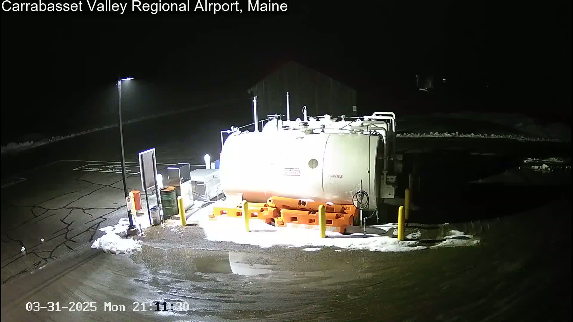 At night, a large white fuel tank sits in a snowy parking lot illuminated by a tall light pole.