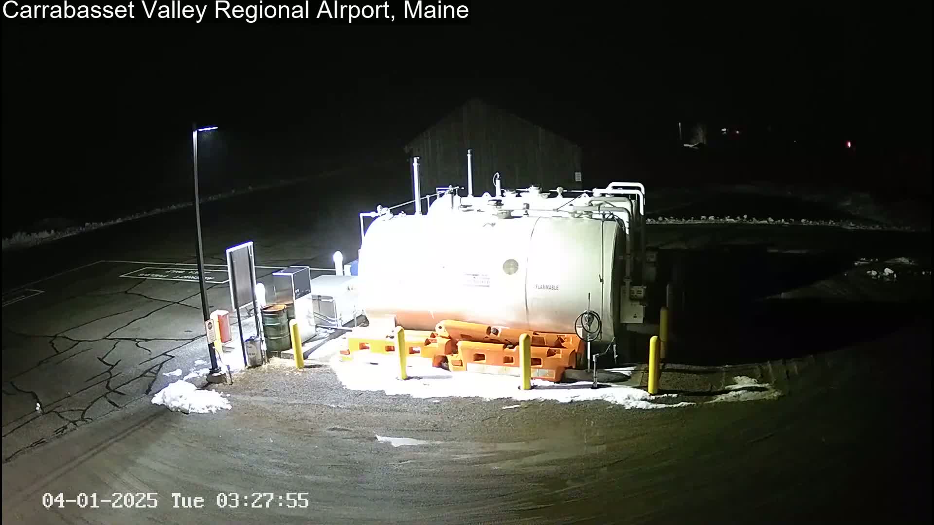 At night, a large, cylindrical fuel tank sits in a dark, snow-dusted parking lot, illuminated by artificial lights.