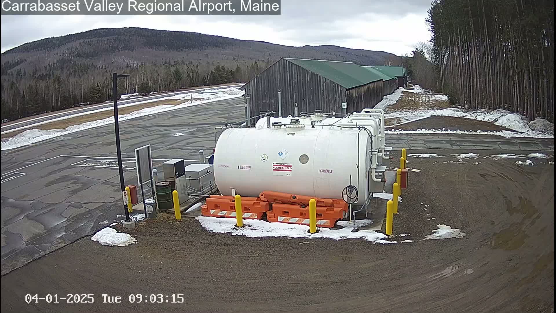 A large white fuel tank sits in a mostly empty asphalt lot with patches of snow, near a long wooden building under a cloudy sky.