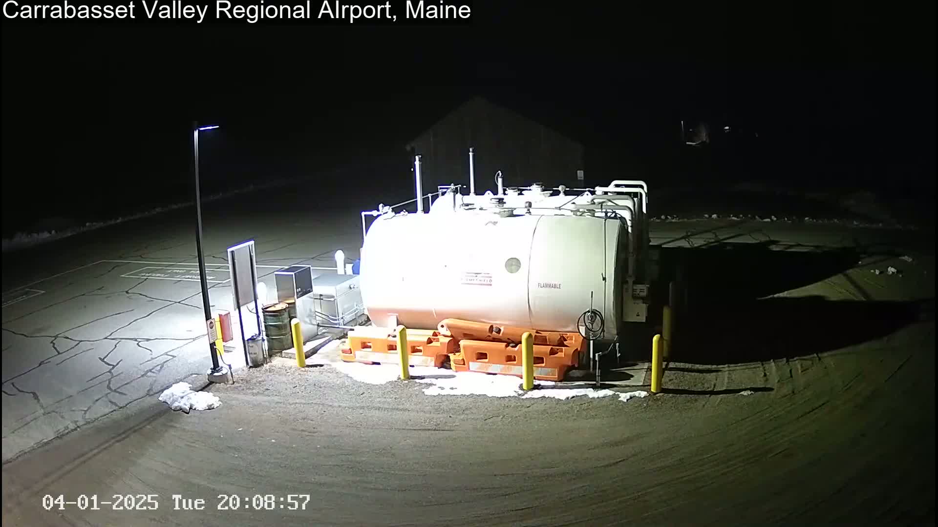 A large, cylindrical fuel tank sits in a dark, nighttime airport parking lot with patches of snow on the ground.