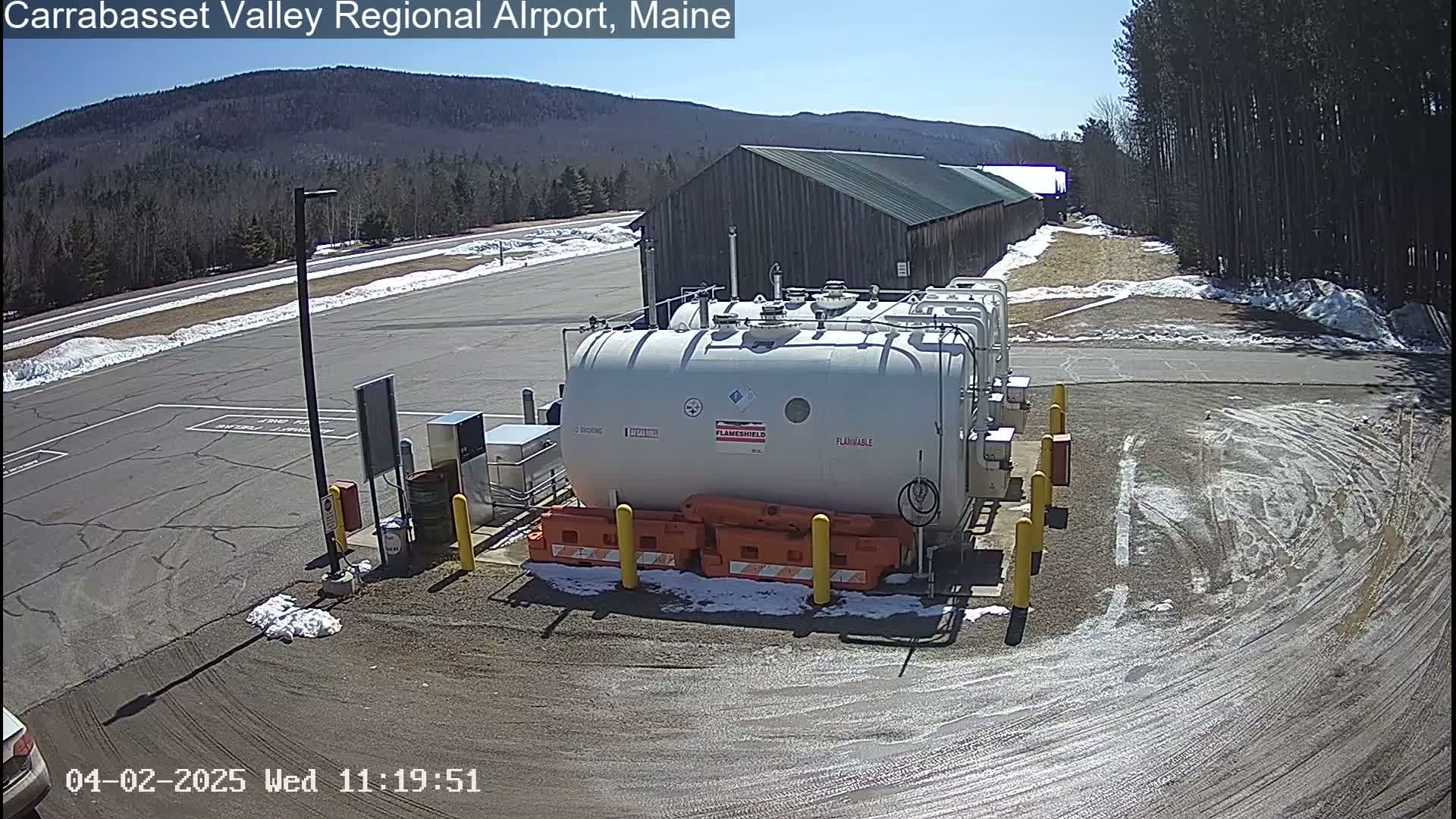 Under a sunny sky, several large white tanks sit in a paved area of an airport, surrounded by snow patches and a long, dark building in the background.
