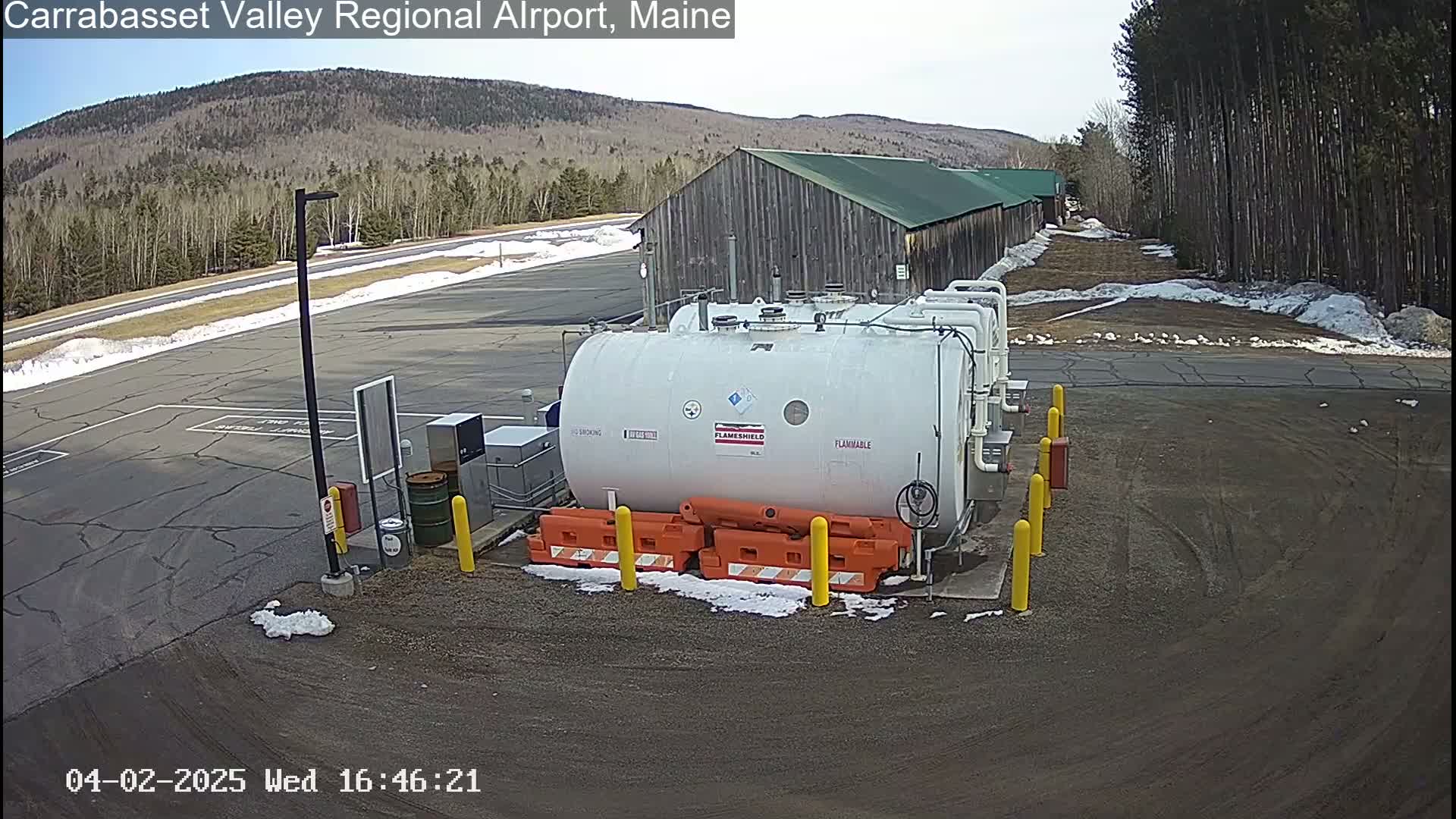 A large white fuel tank sits in a mostly empty asphalt lot next to a long wooden building, under a partly sunny sky with some snow patches on the ground.