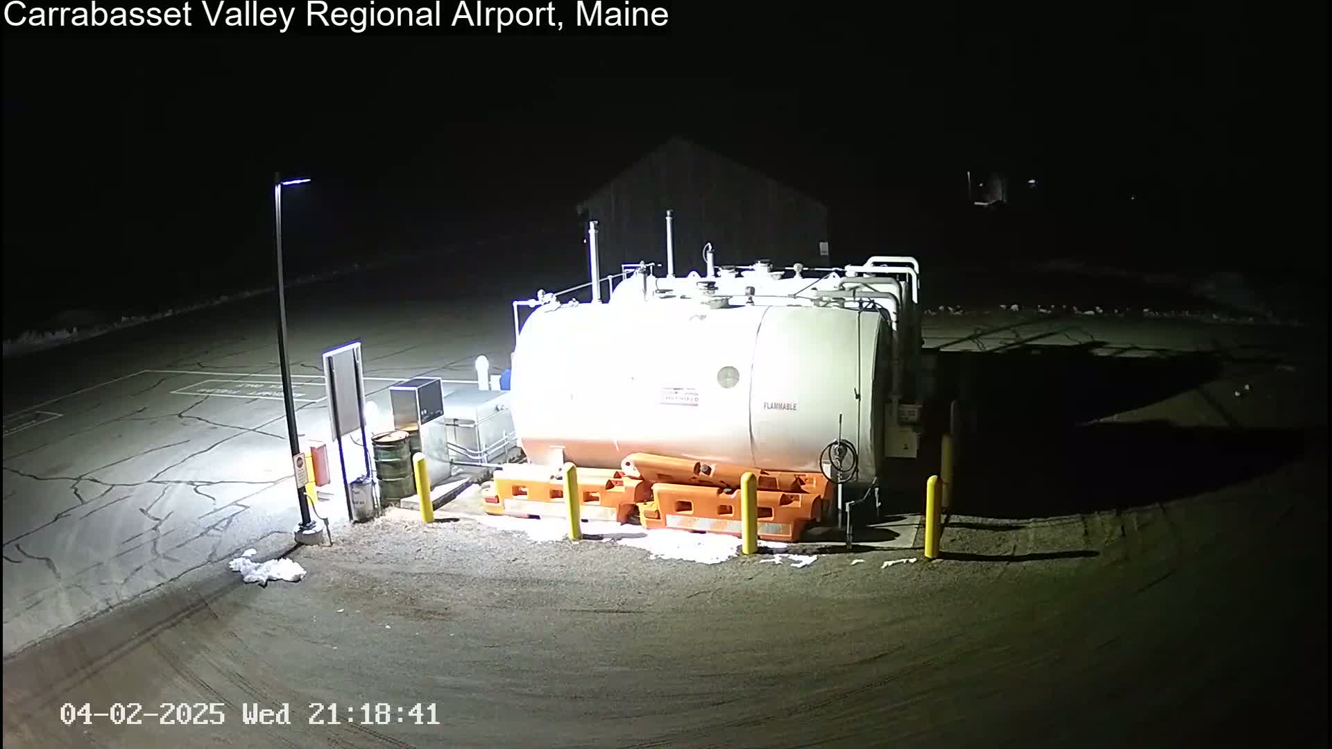 A large, white, flammable fuel tank sits in a dark, empty parking lot at night, illuminated by a single light pole.