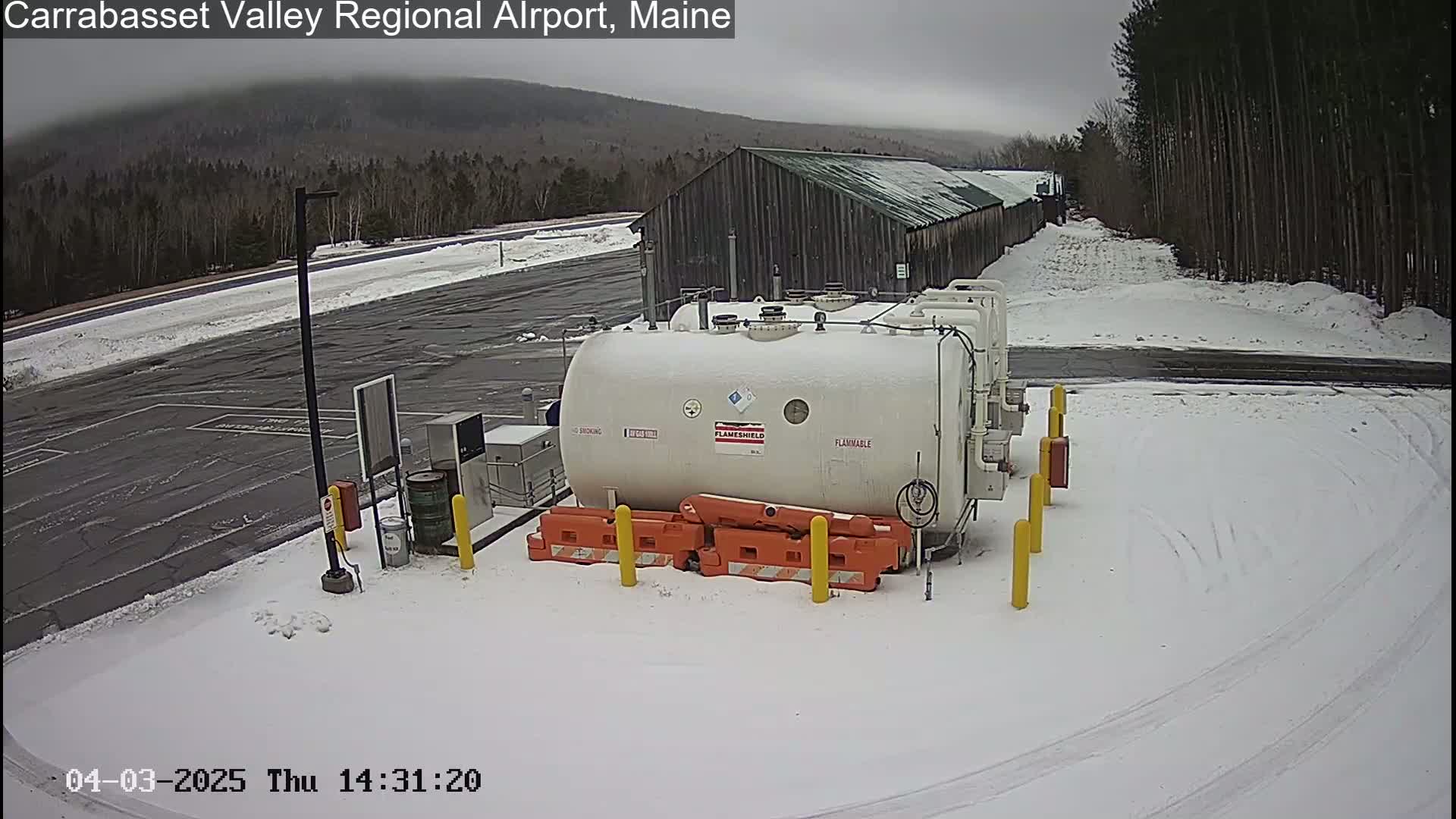 A large white flammable liquid storage tank sits in a snow-covered airport parking lot under an overcast sky, with a dark-colored building and a snow-covered landscape in the background.