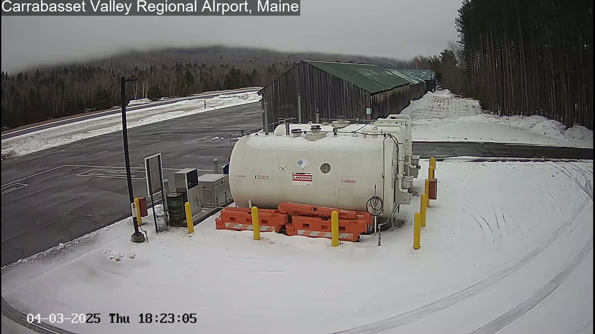 A large white tank labeled "flammable" sits in a snow-covered airport parking lot next to a long, dark-colored building under an overcast sky.