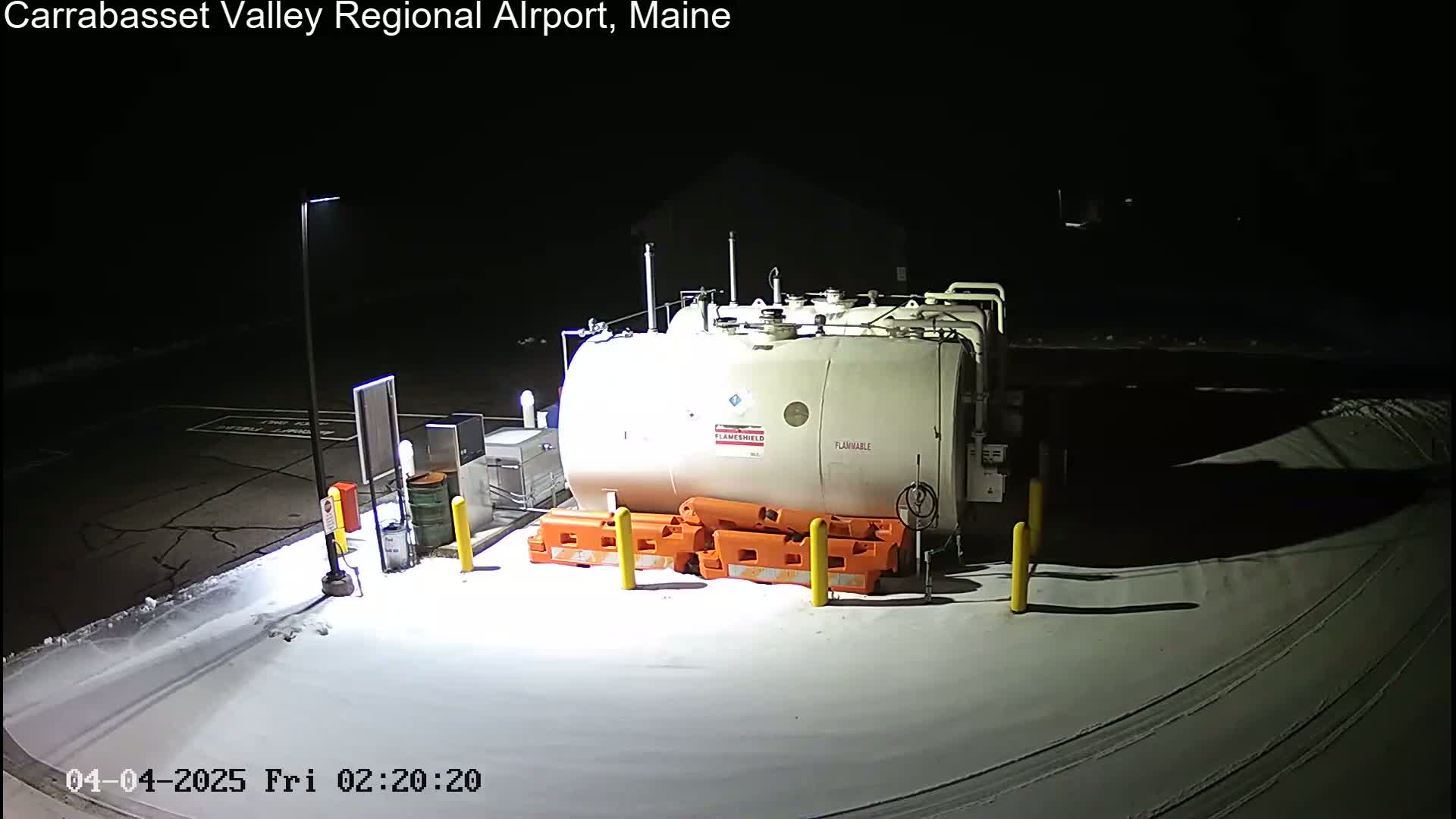 A large, off-white fuel tank sits in a snow-covered area at night, surrounded by orange and yellow safety barriers and illuminated by artificial lights.