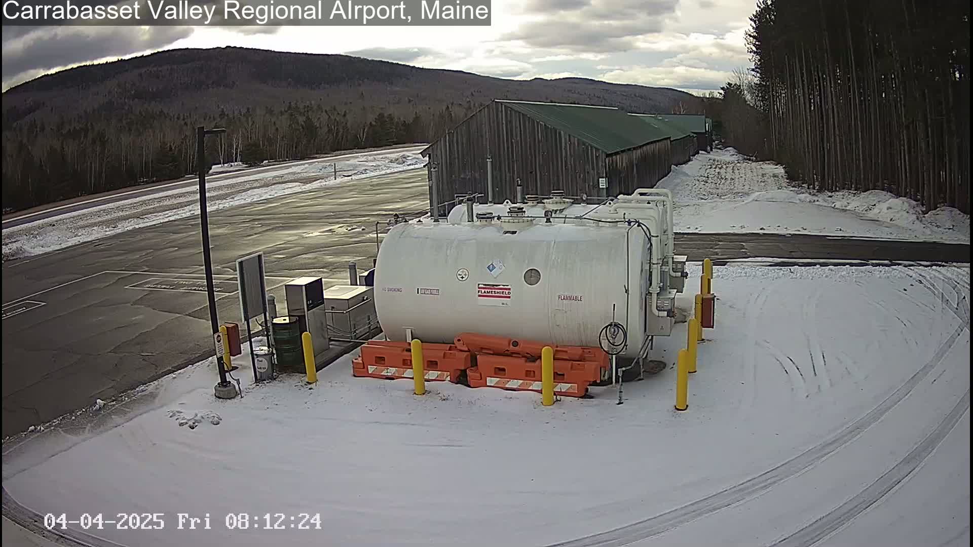 A large white tank sits in a snow-covered airport parking lot near a long, low building, with trees and a mountain visible in the background under a cloudy sky.