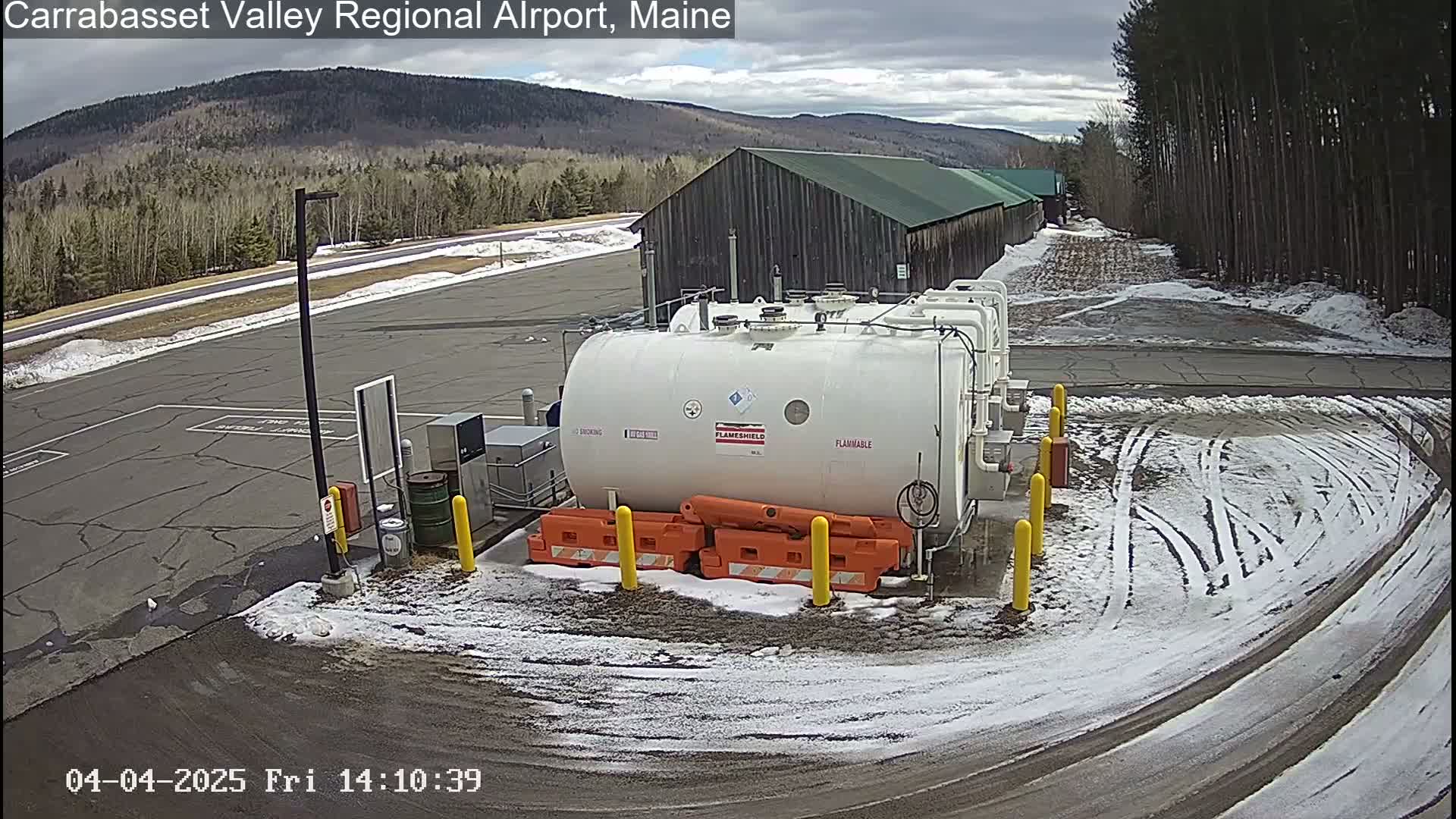 A large white flammable liquid storage tank sits in a snowy airfield parking lot near a long building, with a mountain range visible in the background under an overcast sky.