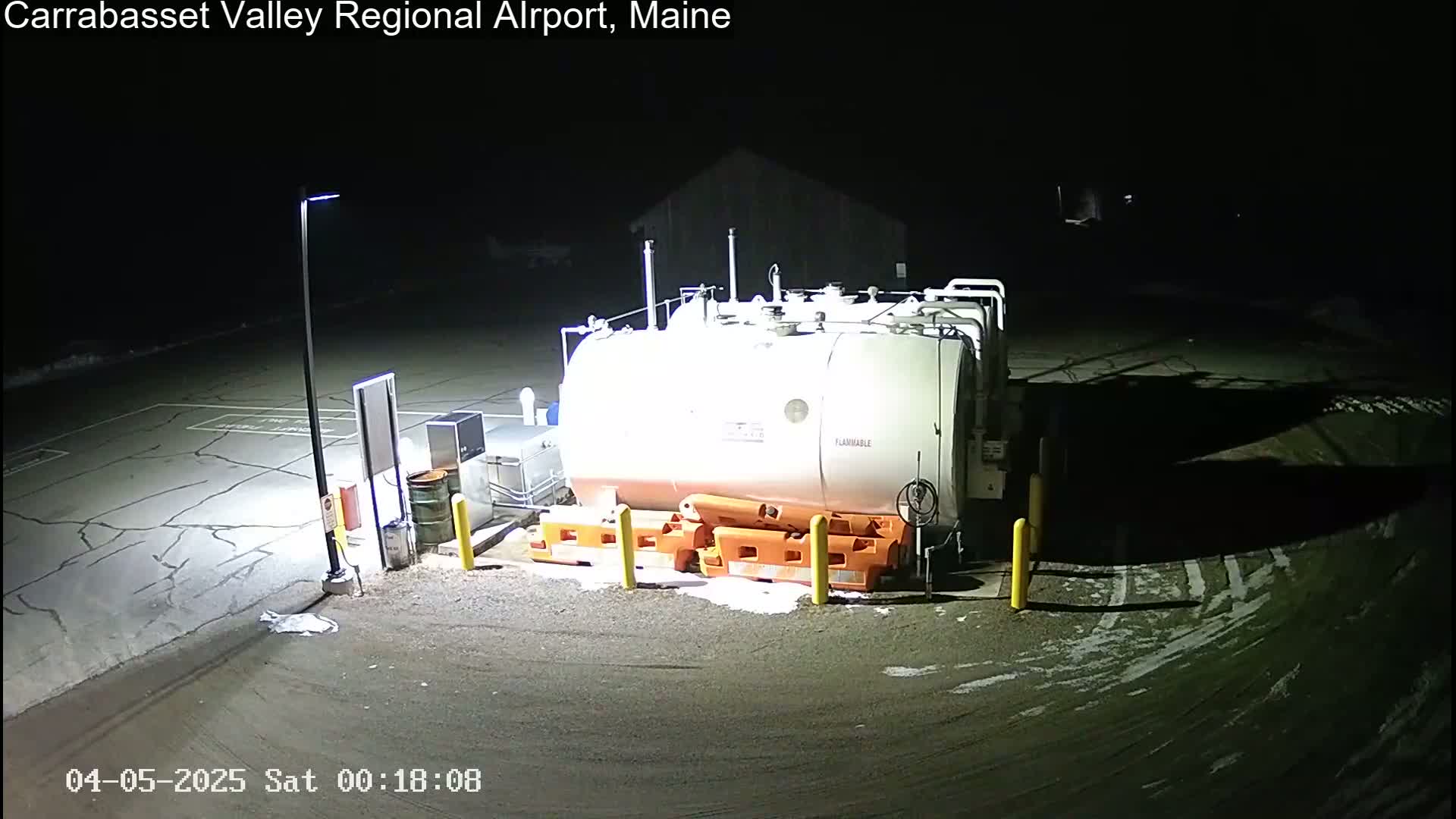 A large, light-colored fuel tank sits in a dark, snow-dusted airport parking lot at night, illuminated by a single lamppost.