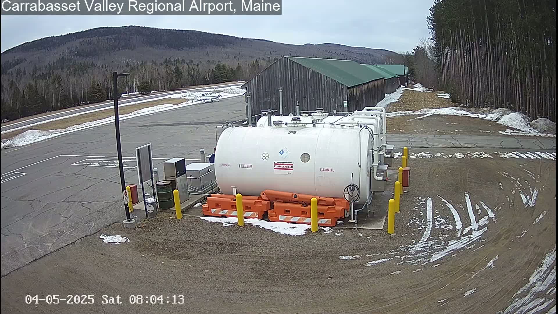 A large white fuel tank sits in a small airport parking lot with patches of snow and tire tracks on the ground, backed by a long building and a distant mountain range under a cloudy sky.