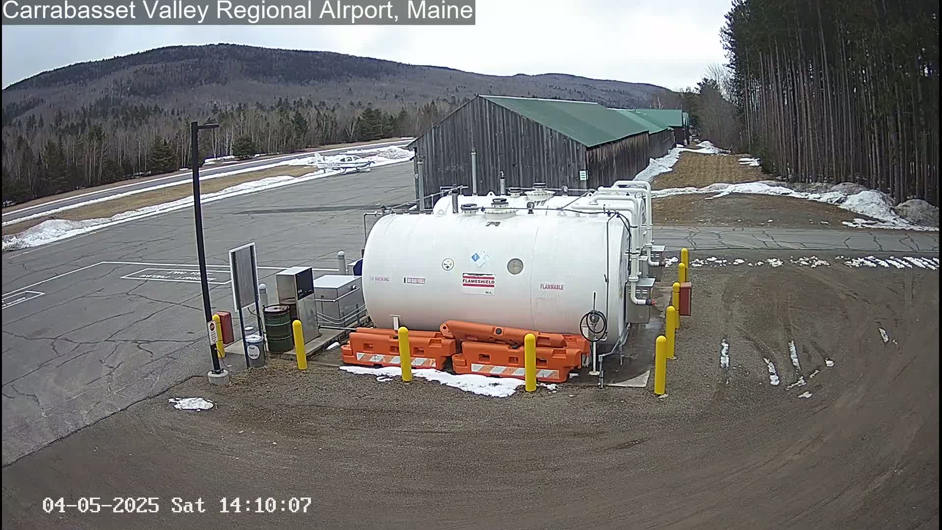 A large white fuel tank sits in a paved area of an airport, with a small plane visible in the background under an overcast sky.