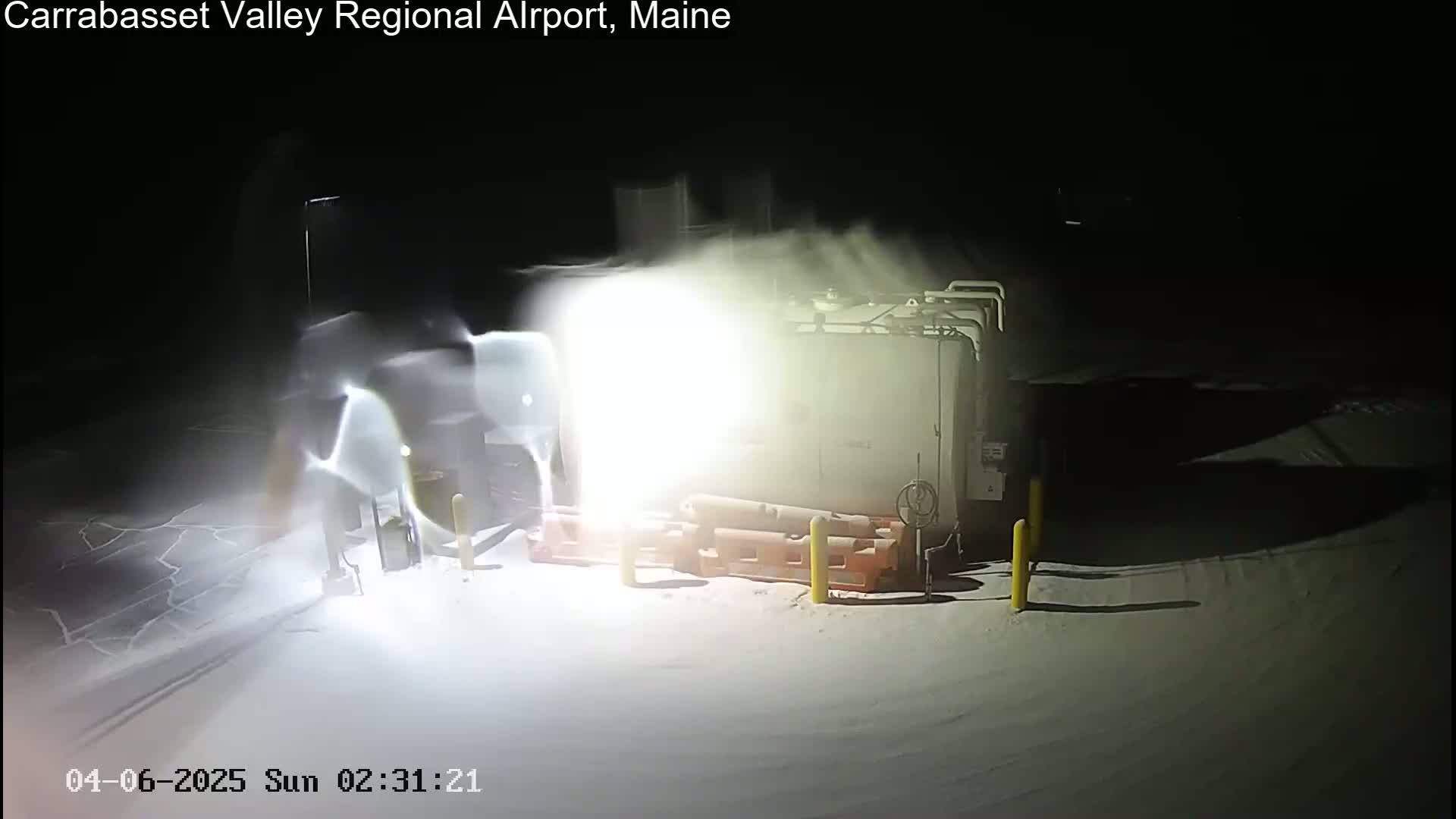 A bright light illuminates a snow-covered area at night near a large tank and some equipment, with blowing snow obscuring some details.