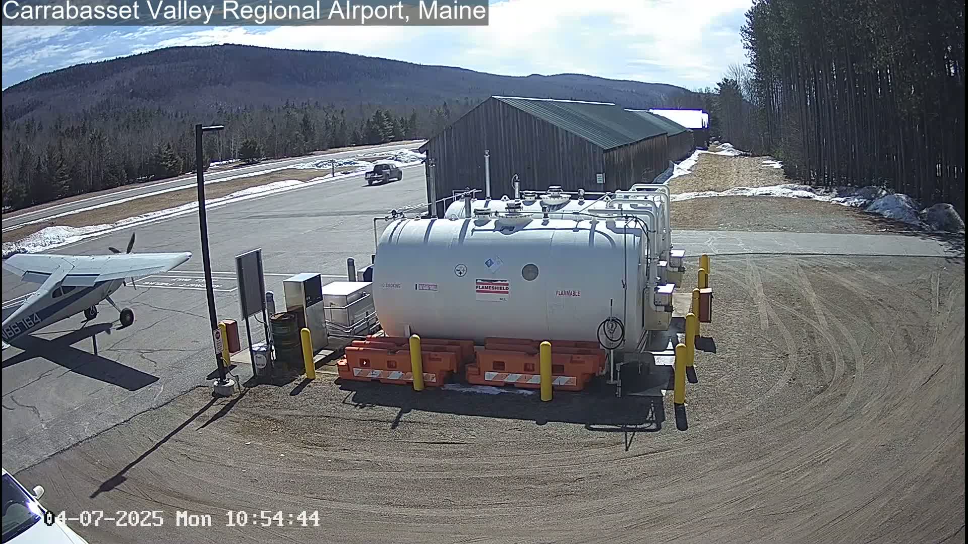 A small airplane is parked near a fuel storage facility at an airport on a sunny day with some snow on the ground.
